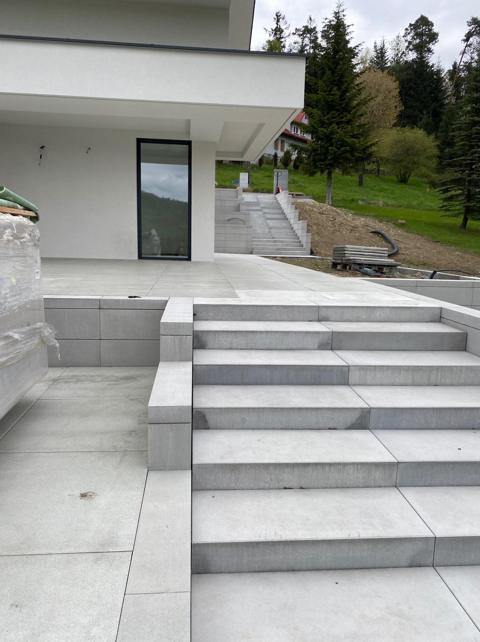 Nowoczesne schody zewnętrzne z jasnoszarej kostki brukowej prowadzące do domu z minimalistyczną architekturą, zieleń w tle. Widoczne narzędzia i materiały budowlane.