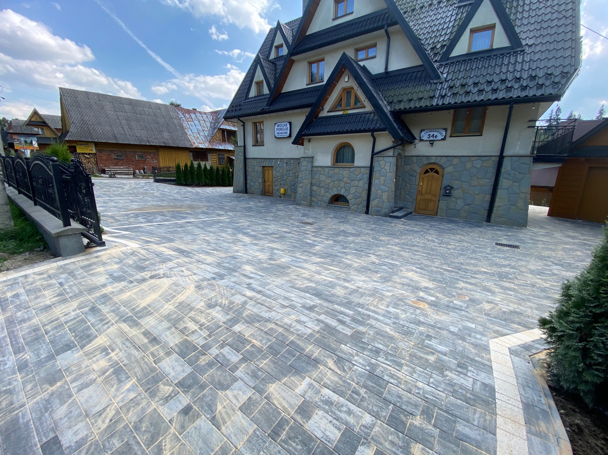 Szara kostka brukowa ułożona przed budynkiem w stylu góralskim, z widocznym ogrodzeniem i elementami zieleni. Jasne niebo z delikatnymi chmurami w tle.