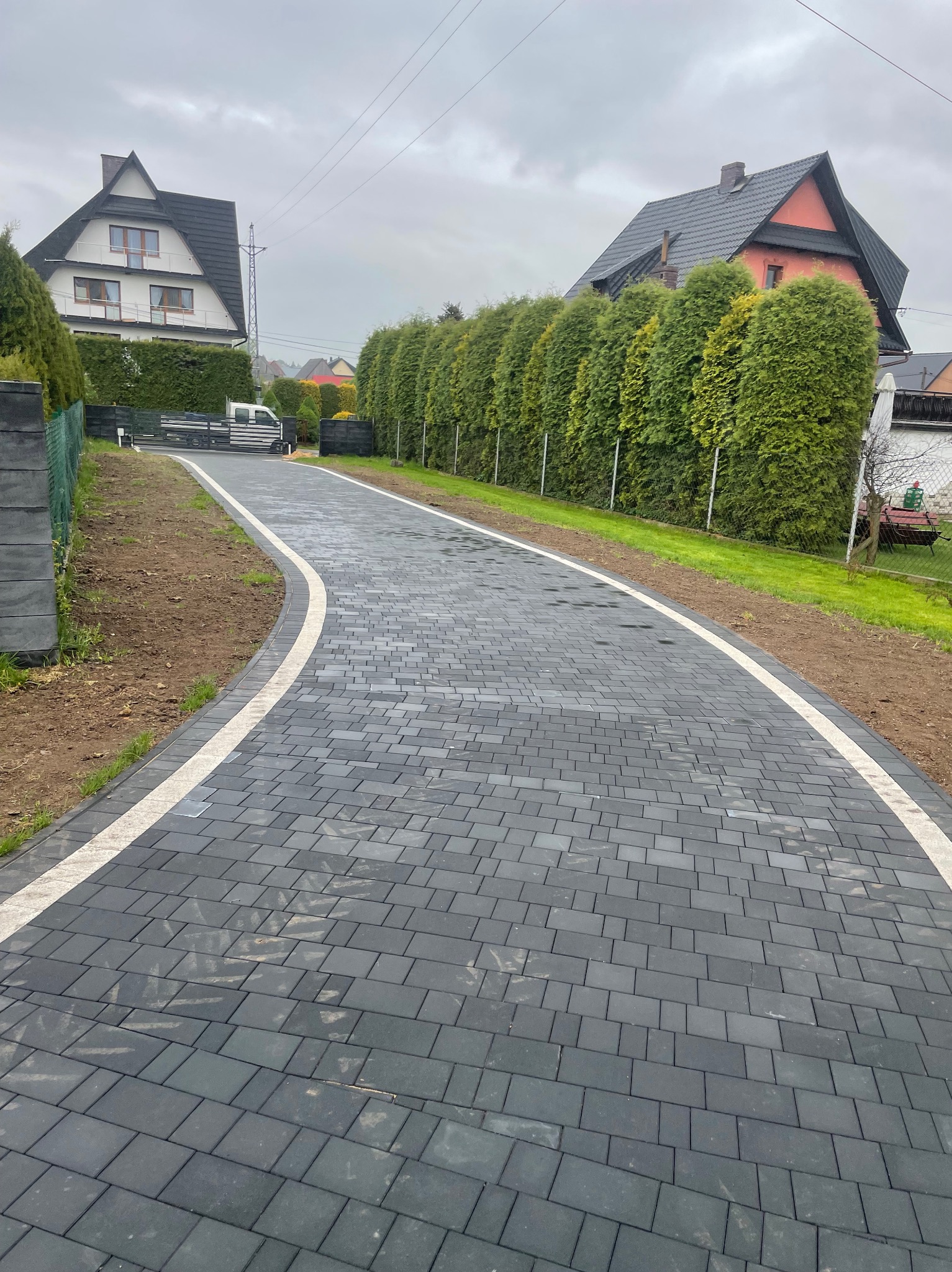 Czarna kostka brukowa z białym obramowaniem tworzy elegancki podjazd na posesję. W tle zadbane tuje i domy.
