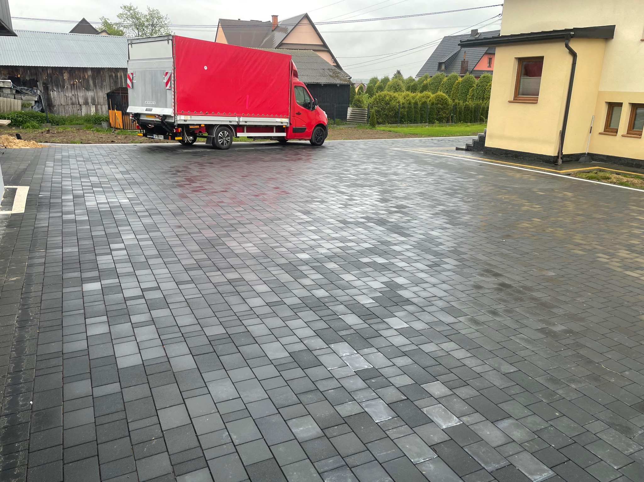 Ułożona, szara kostka brukowa na posesji z zaparkowanym czerwonym samochodem dostawczym. Widoczne budynki w tle i świeżo ułożone obrzeża.
