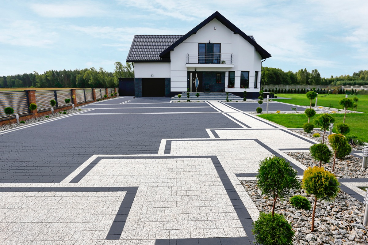 Dwukolorowa kostka brukowa ułożona we wzór geometryczny przed nowoczesnym domem z białymi ścianami i ciemną dachówką, ozdobne krzewy kuliste wzdłuż ścieżki.
