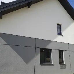 Nowoczesny dom z szarą elewacją, trzema oknami z ciemnymi ramami i skrzynkami gazowymi na ścianie, widok z zewnątrz w słoneczny dzień.