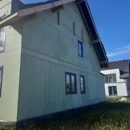 Surowy stan budynku w trakcie budowy z zamontowanymi oknami w ciemnych ramach i zielonymi ścianami zewnętrznymi, widok z zewnątrz na tle błękitnego nieba.