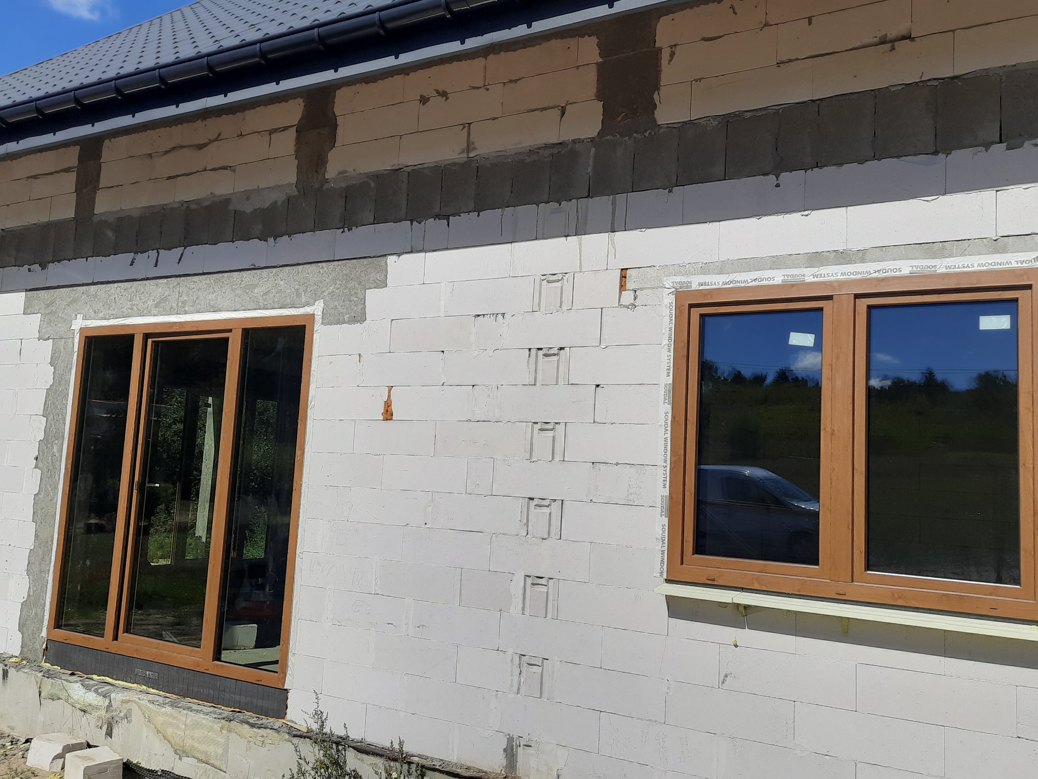 Montaż okien PCV z brązowymi ramami w surowym stanie budynku z białych bloczków, widoczne zabezpieczenia Soudal Window System.