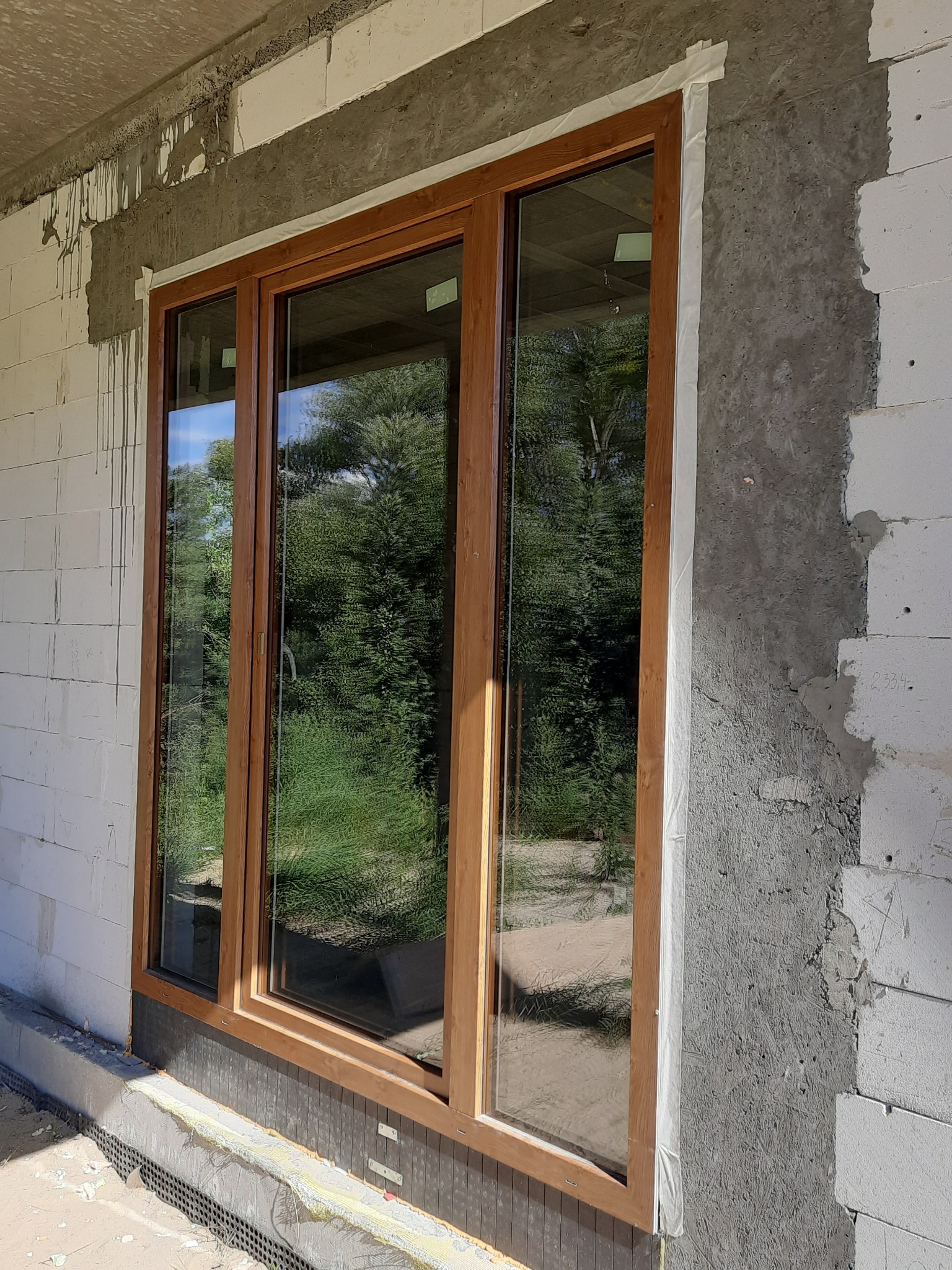 Montaż okna z drewnopodobną ramą w surowym stanie budynku, widoczne bloczki betonowe i świeży tynk, zabezpieczone taśmą malarską.