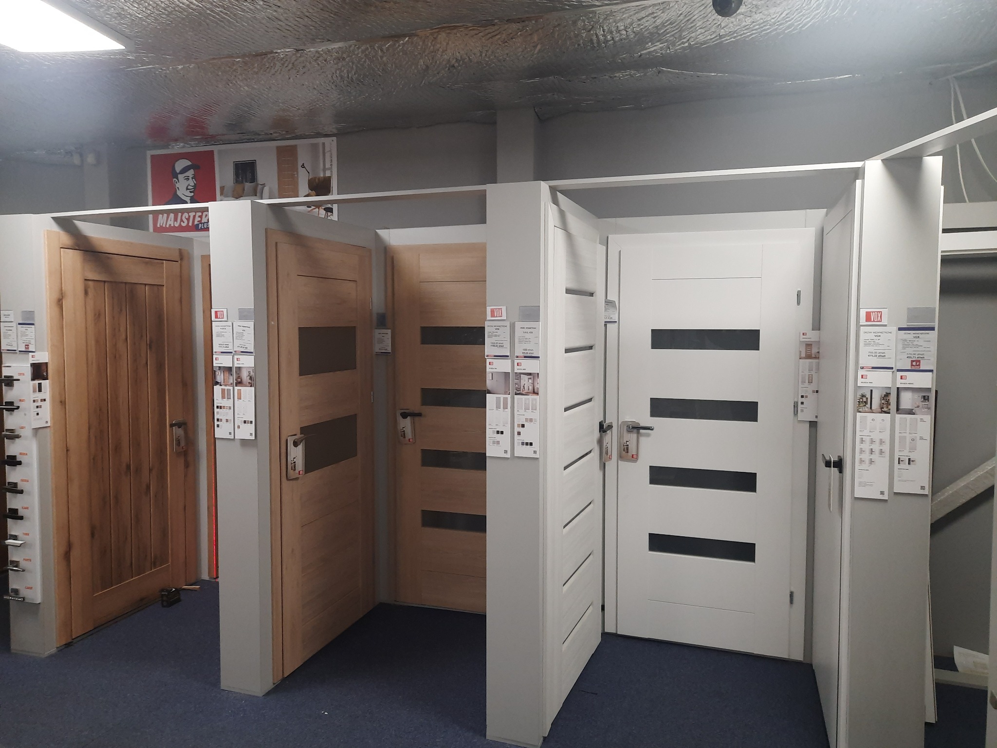 Ekspozycja różnych modeli drzwi wewnętrznych w sklepie, widoczne drzwi drewniane i białe z poziomymi przeszkleniami, na drzwiach zawieszone są ulotki informacyjne.