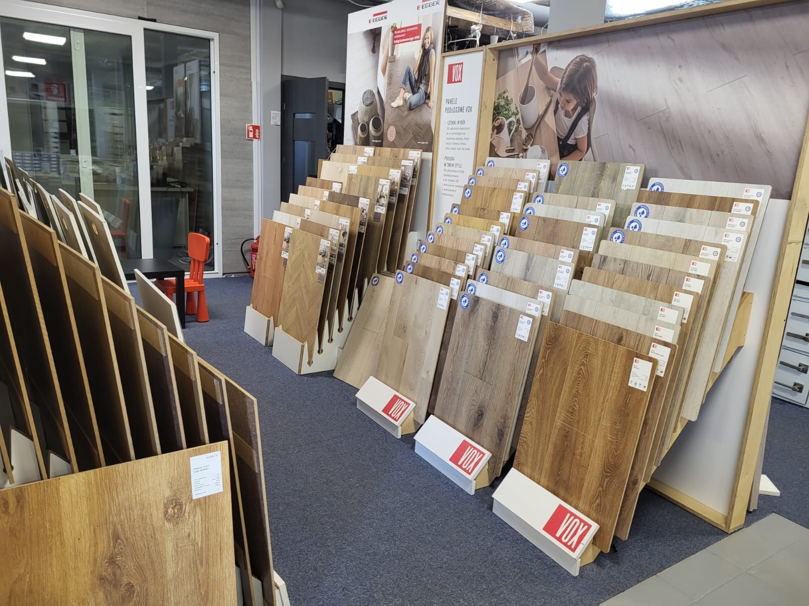 Ekspozycja paneli podłogowych VOX w sklepie, różne odcienie drewna, wzory i faktury, widoczne materiały reklamowe producenta.