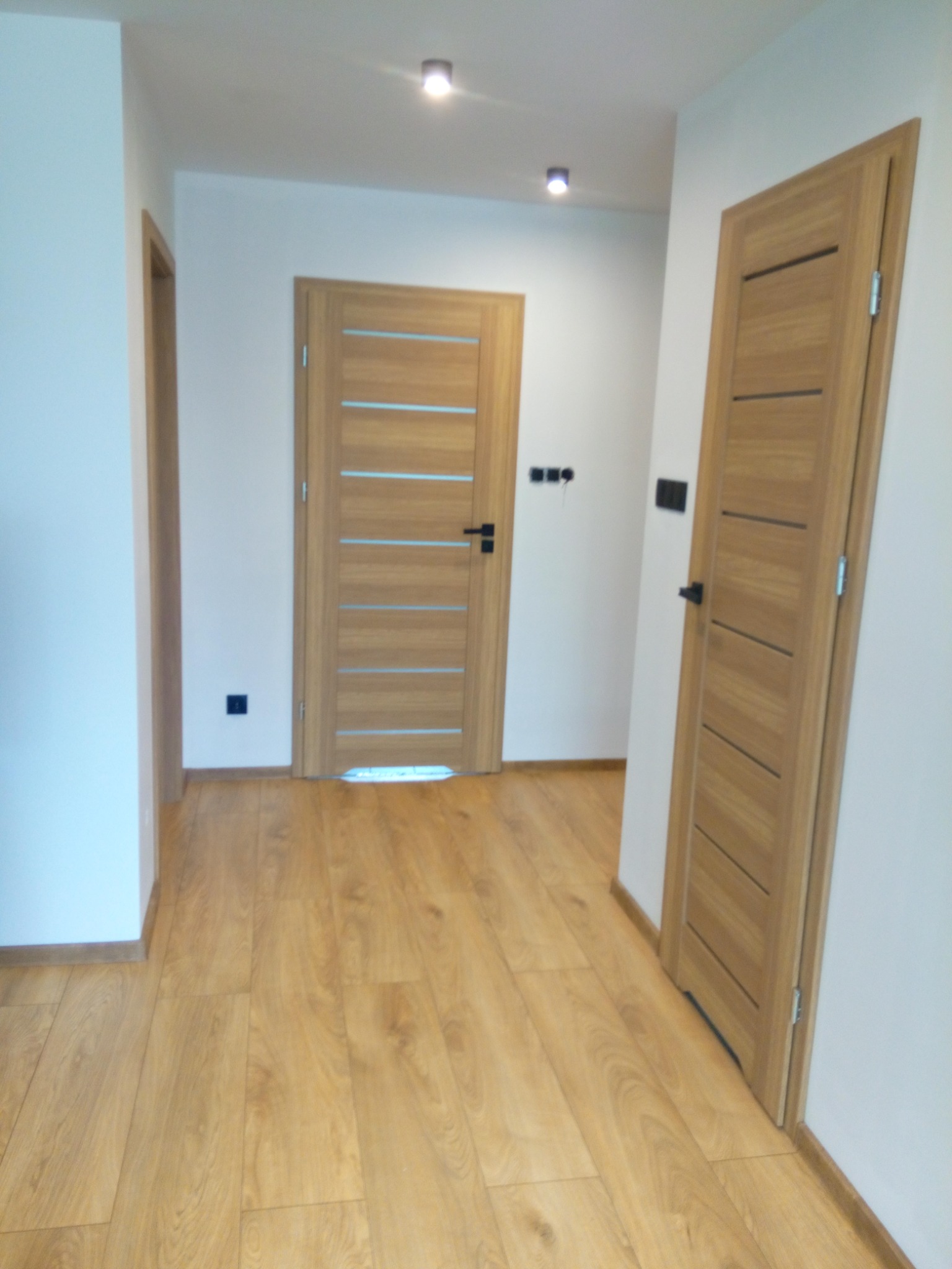 Dwa skrzydła drzwiowe wewnętrzne z poziomymi przeszkleniami w odcieniach drewna, zamontowane w jasnym korytarzu z panelami podłogowymi w podobnej kolorystyce.