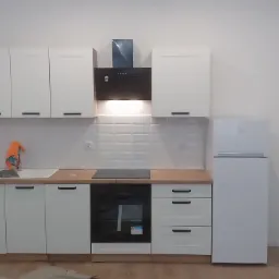 Nowoczesna, biała zabudowa kuchenna z drewnianym blatem, czarnym okapem i płytą indukcyjną, uzupełniona białą lodówką, widoczna na tle białych płytek i ścian.