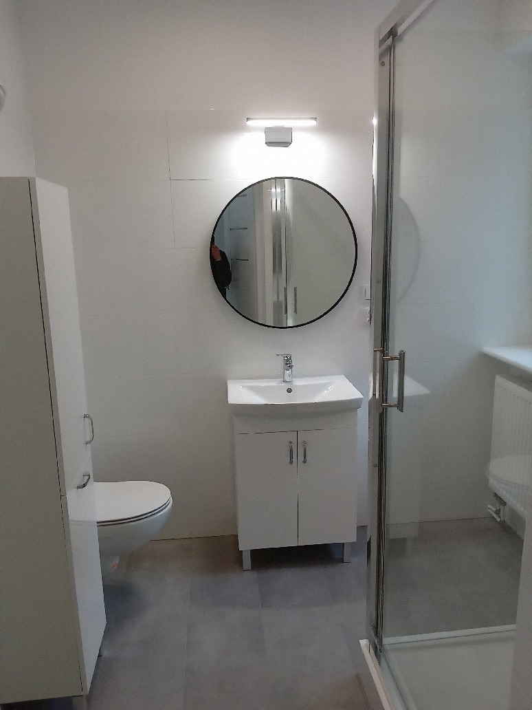 Wykończona łazienka w bieli i szarościach z okrągłym lustrem i kabiną prysznicową, widoczne płytki na ścianach i podłodze, minimalistyczny styl.