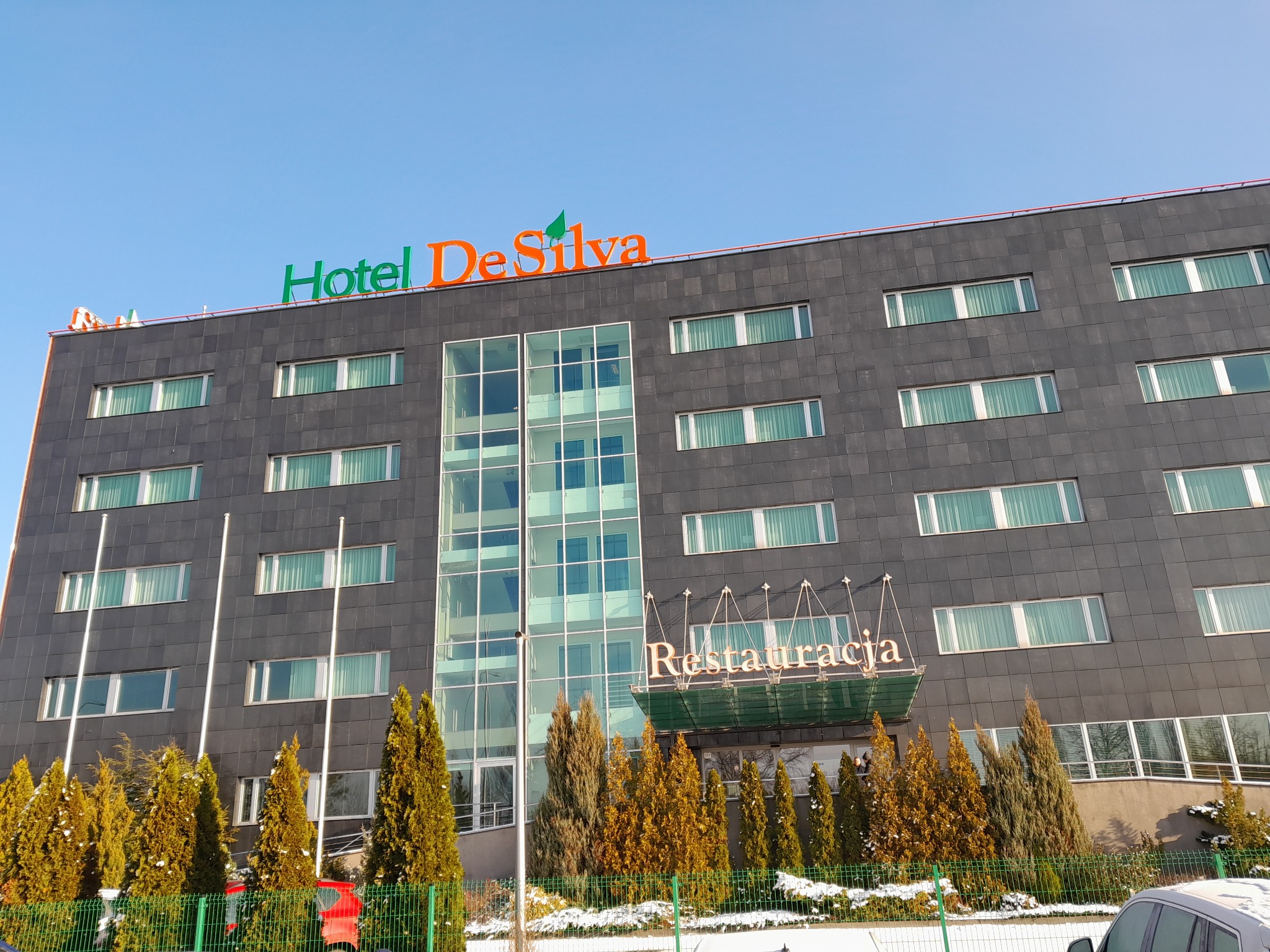 Zewnętrzny widok nowoczesnego hotelu z ciemną elewacją, zielonym i pomarańczowym logo 'Hotel DeSilva' na dachu oraz napisem 'Restauracja' nad wejściem, otoczonego zimozielonymi krzewami z resztkami...