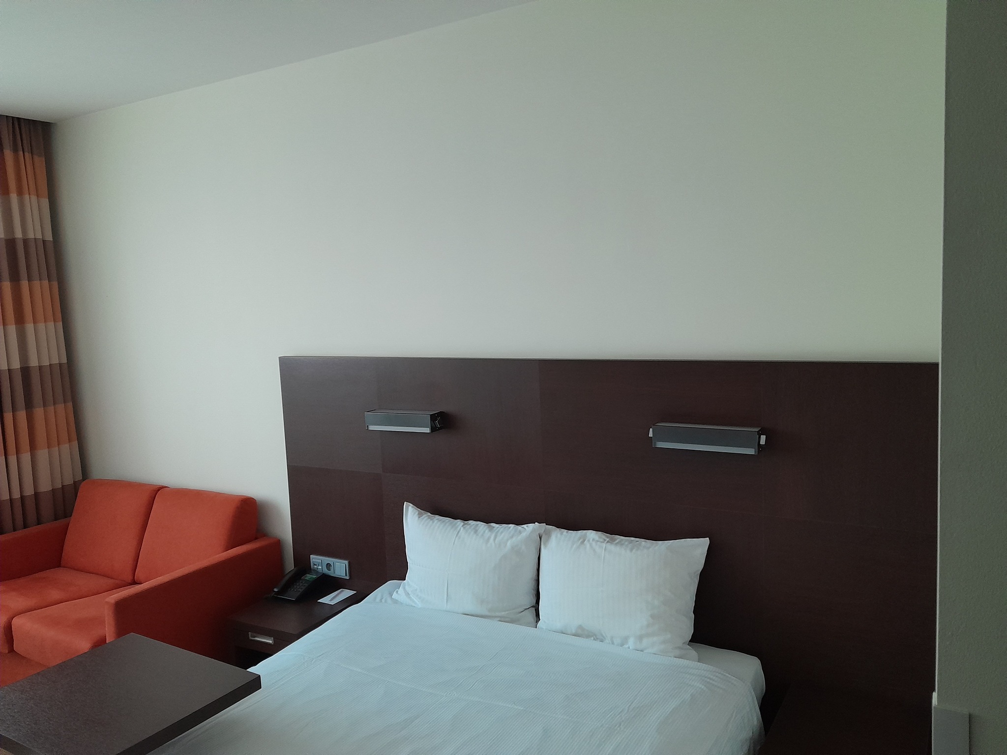 Pokój hotelowy z pomarańczową sofą, łóżkiem z białą pościelą i stolikiem nocnym z telefonem, oświetlony lampami na wezgłowiu.