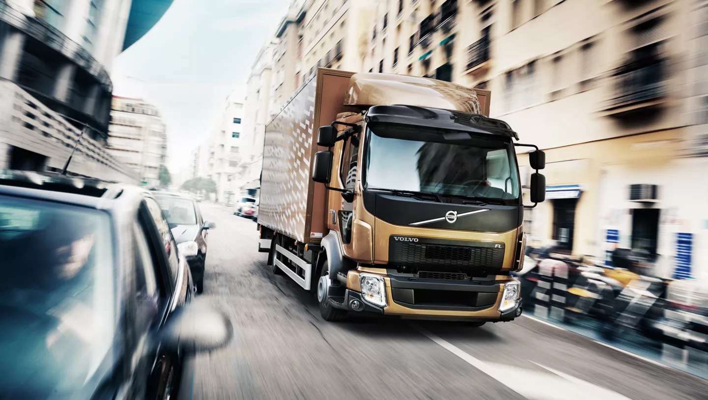 Złoty samochód ciężarowy Volvo FL w ruchu ulicznym, z rozmytym tłem budynków miejskich i innymi pojazdami, dynamiczne ujęcie.