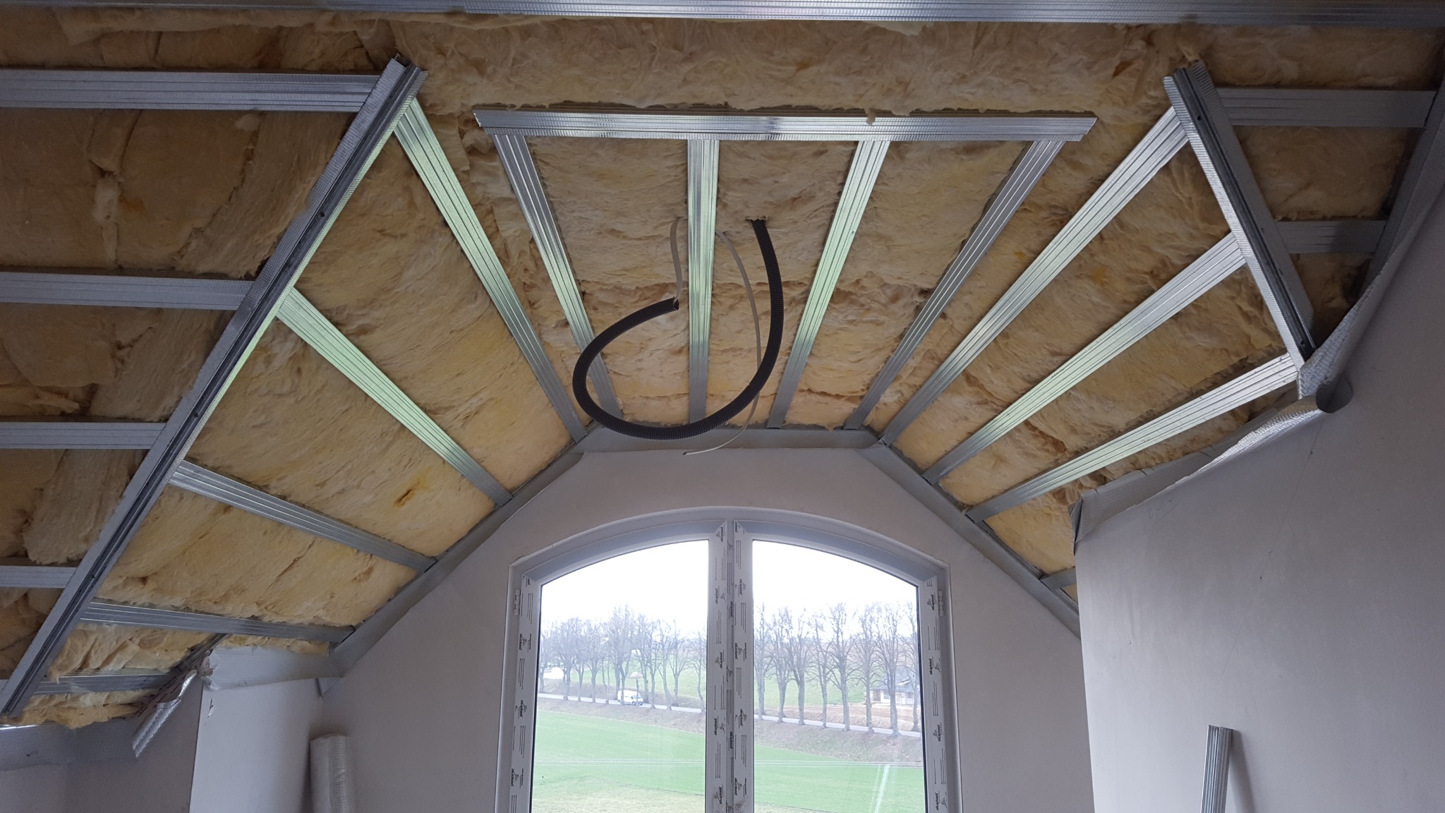 Montaż izolacji z wełny mineralnej w poddaszu, widoczne metalowe profile sufitowe, okno dachowe z widokiem na zielone pole i drzewa.