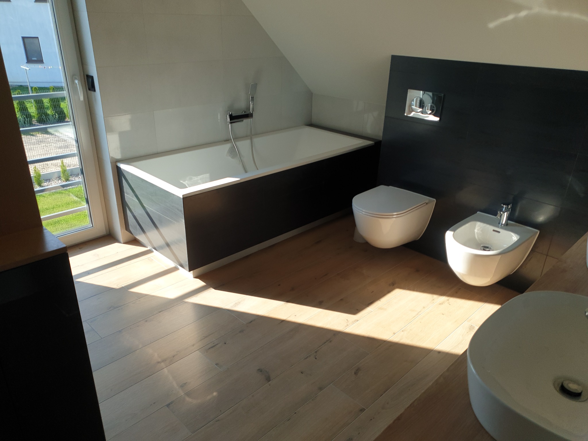 Nowoczesna łazienka na poddaszu z wanną, toaletą podwieszaną i bidetem, wykończona białymi i grafitowymi płytkami oraz drewnianą podłogą, oświetlona promieniami słońca wpadającymi przez okno.