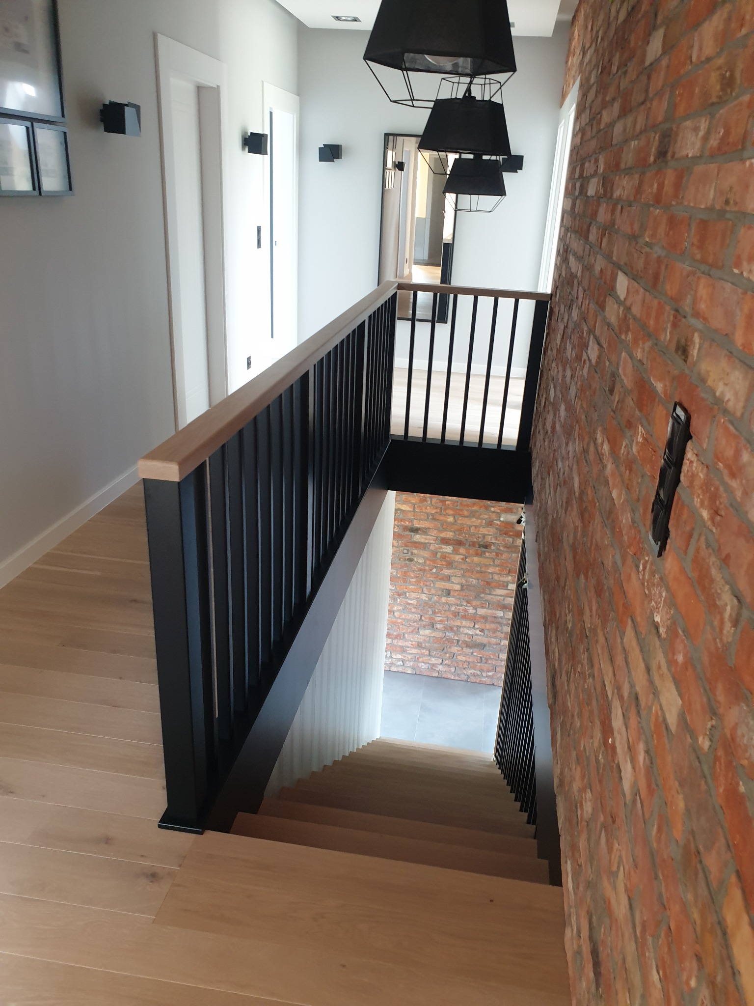 Nowoczesne schody z drewnianymi stopniami i czarną balustradą, ściana wyłożona czerwoną cegłą, widok z góry na dół, oświetlenie ledowe.
