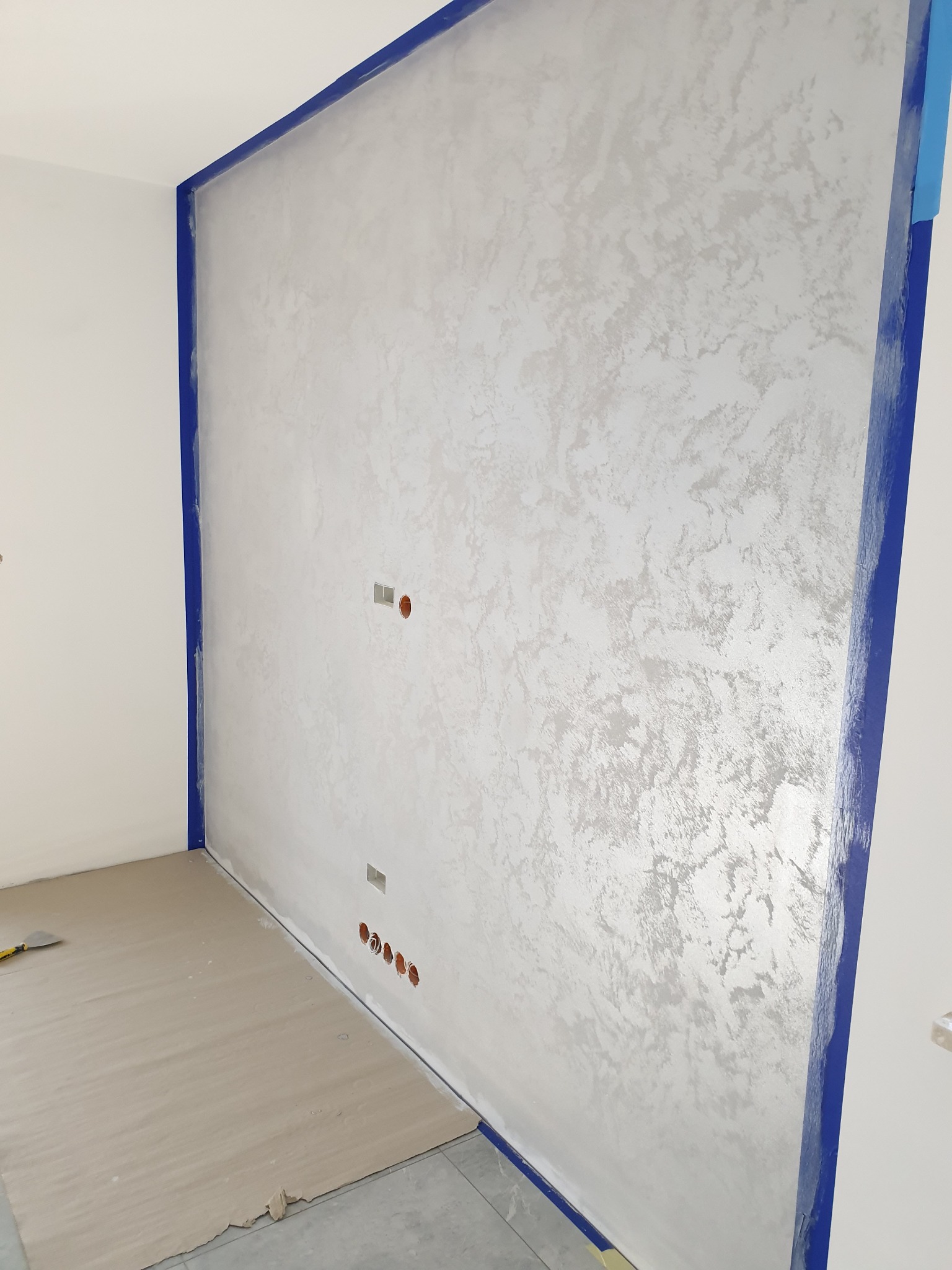 Ściana w pokoju po nałożeniu dekoracyjnej, srebrzystej farby strukturalnej, zabezpieczona niebieską taśmą malarską, widoczne gniazdka elektryczne i zabezpieczona podłoga.