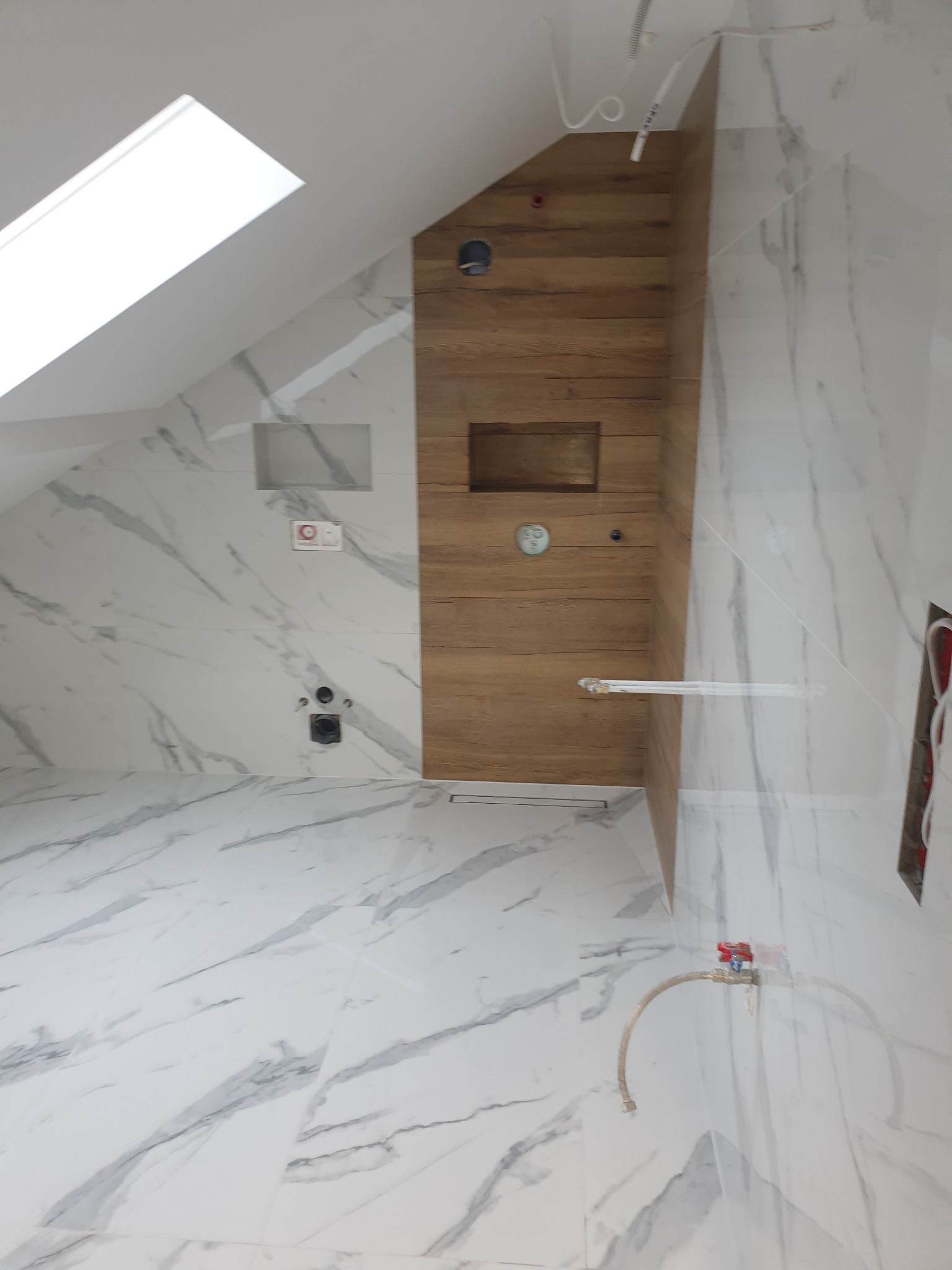 Wykończona łazienka na poddaszu ze skośnym oknem dachowym, marmurowe płytki podłogowe i ścienne z szarymi żyłami, drewnopodobna zabudowa prysznica z półkami i deszczownicą, widoczne przyłącza wodne.