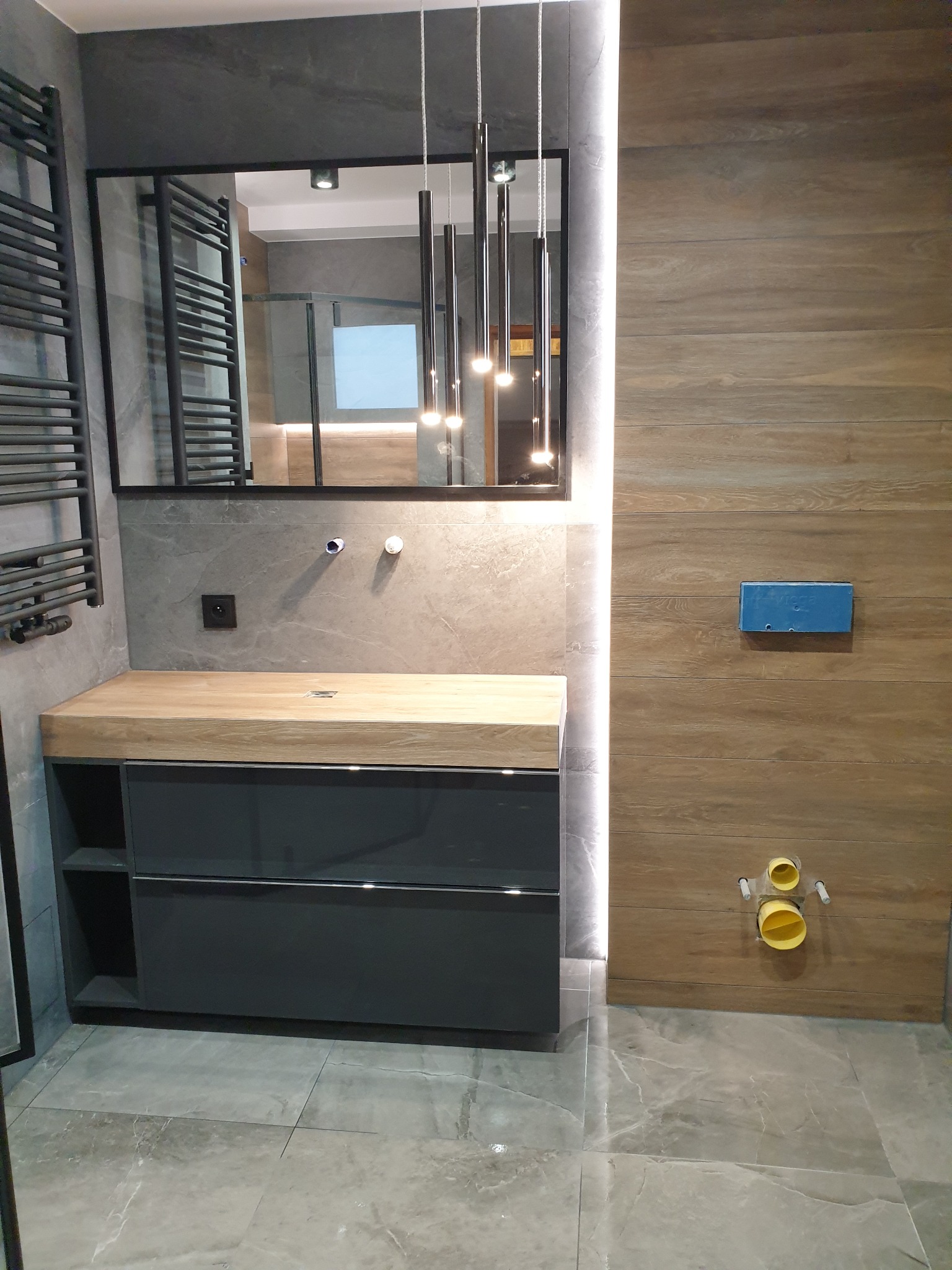 Nowoczesna łazienka z szarymi płytkami imitującymi kamień, drewnianą szafką pod umywalką z szufladami i lustrem w czarnej ramie, oświetlona wiszącymi lampami.