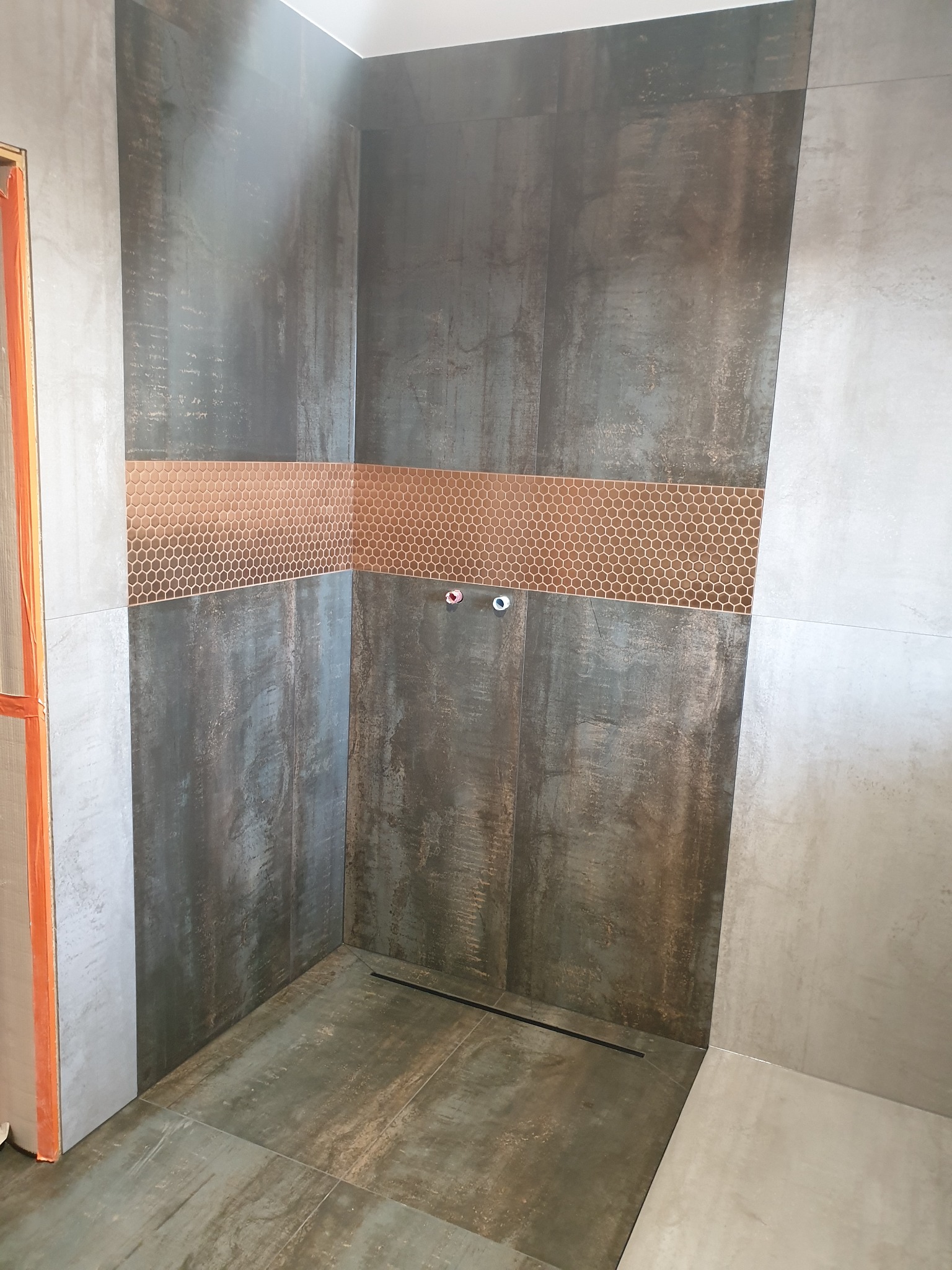 Wykończona kabina prysznicowa z ciemnymi, metalizowanymi płytkami i poziomym pasem mozaiki w odcieniach miedzi. Widoczny odpływ liniowy w podłodze i wystające końcówki instalacji wodnej.