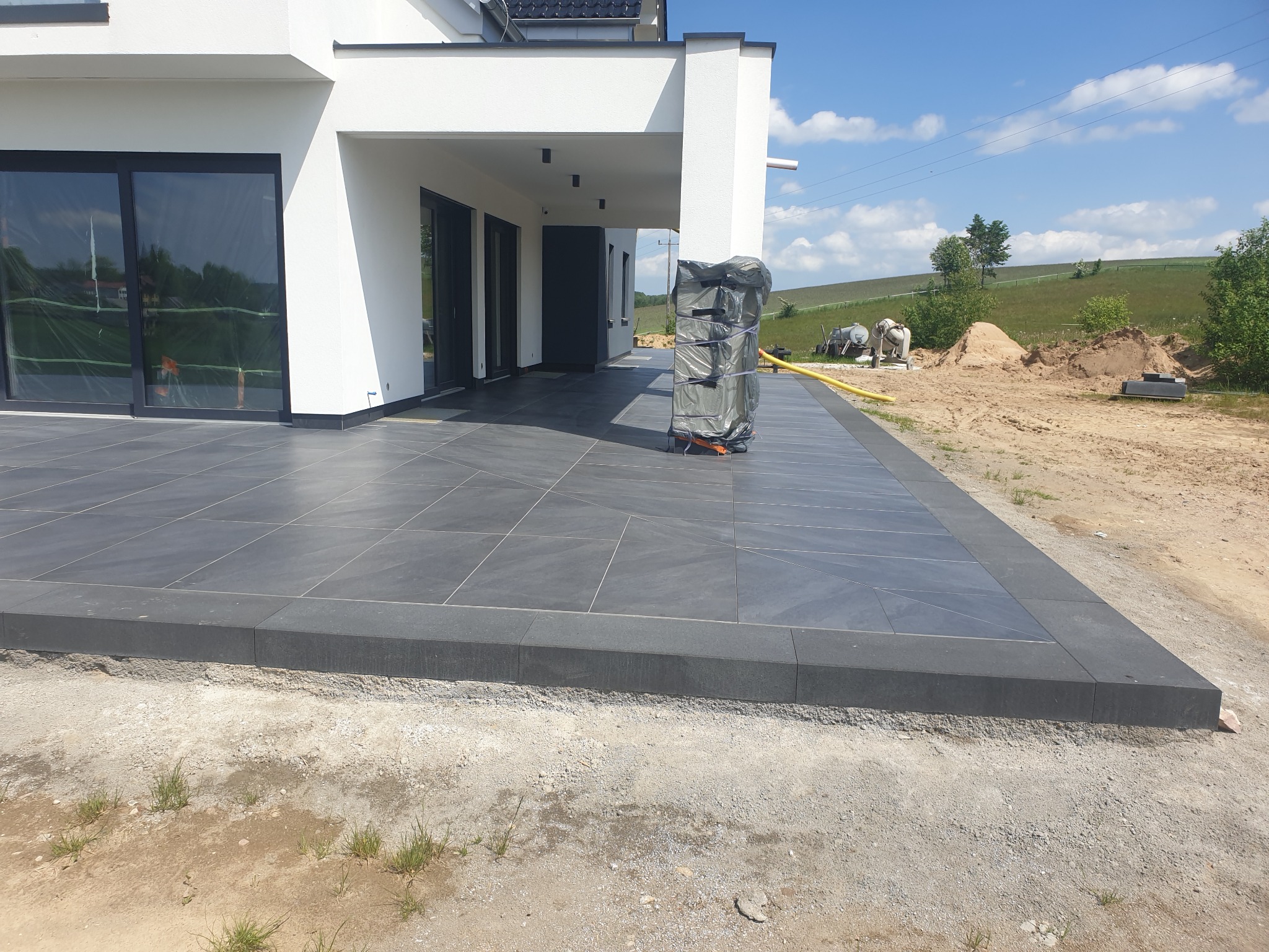Nowoczesny taras z szarych, kwadratowych płytek ceramicznych, obrzeża z ciemniejszego kamienia, widok na biały dom i krajobraz w tle, częściowo zakończone prace ziemne.