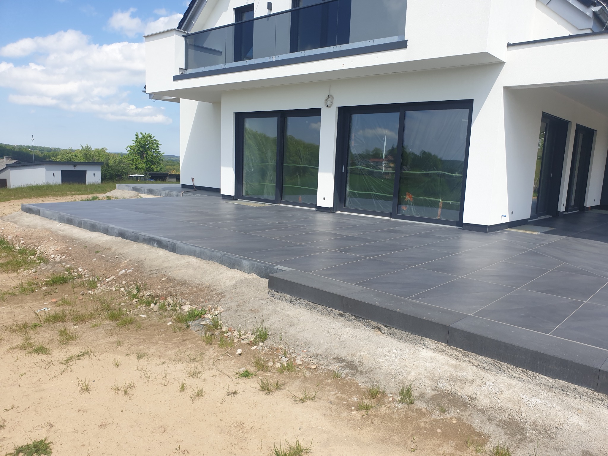 Nowoczesny taras z ciemnoszarej, kwadratowej terakoty, z widocznymi fugami, przylegający do białego budynku z dużymi, przeszklonymi drzwiami przesuwnymi, częściowo zabezpieczonymi folią. Widoczne...