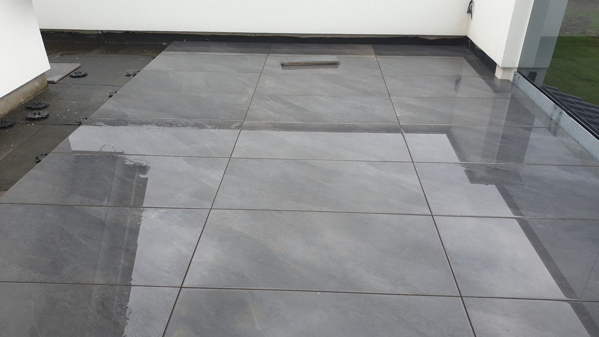 Układanie szarych płytek gresowych na tarasie z widocznymi dystansami poziomującymi i częściowo mokrą powierzchnią odbijającą otoczenie.