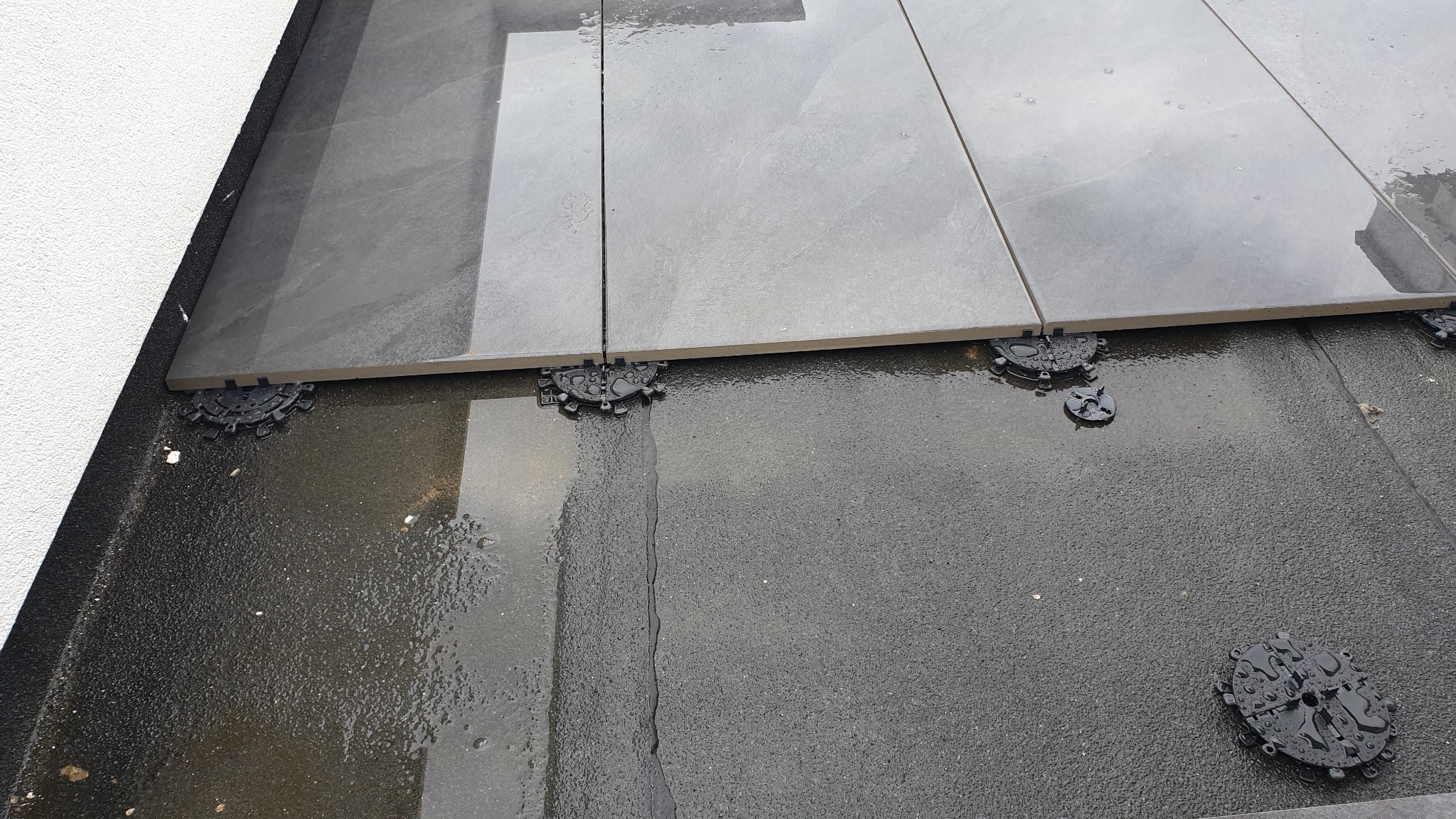 Montaż szarych płyt tarasowych na regulowanych wspornikach z tworzywa sztucznego na mokrym podłożu, widoczna ściana budynku w tle.