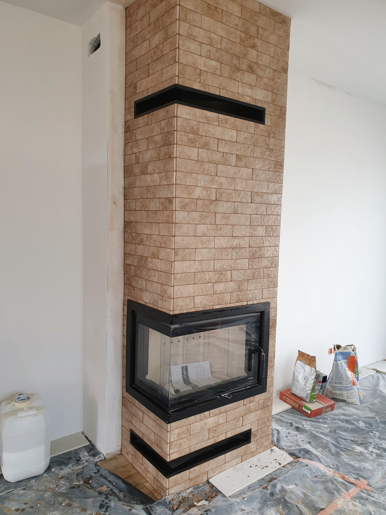 Obudowa kominka wykończona beżowymi płytkami imitującymi cegłę, z czarnymi poziomymi wstawkami i czarną ramą wkładu kominkowego. Podłoga zabezpieczona folią, widoczne worki z klejem i pojemnik...