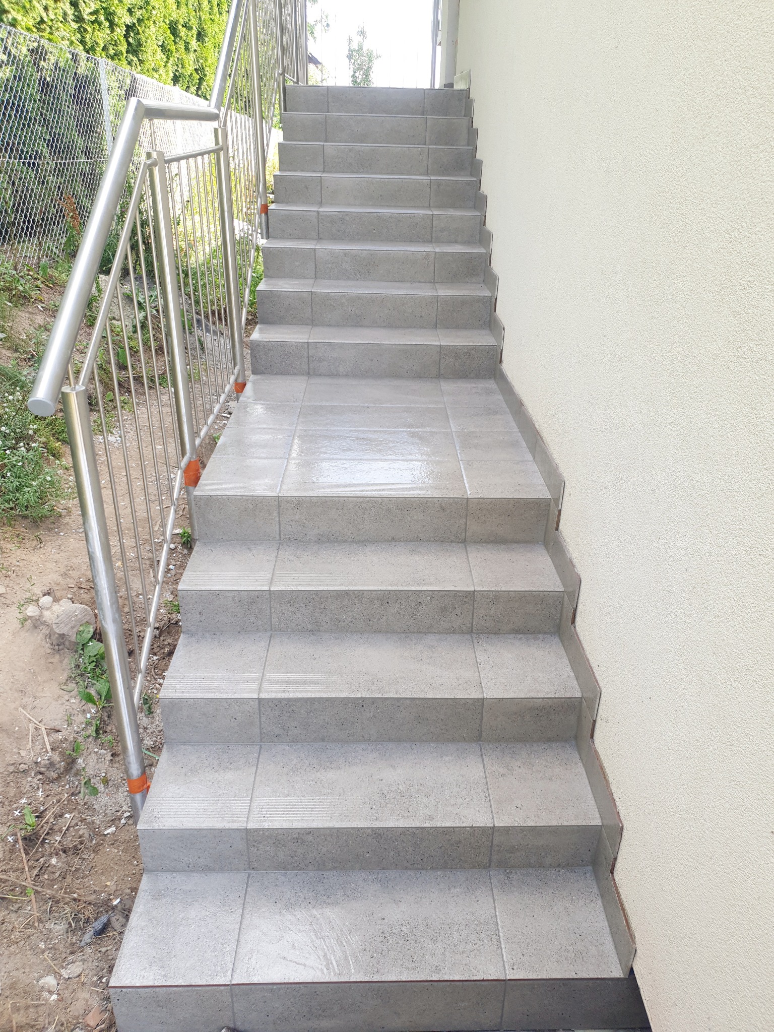 Zewnętrzne schody wyłożone szarymi płytkami z metalową barierką po lewej stronie, prowadzące do góry, z widoczną ścianą budynku po prawej stronie.