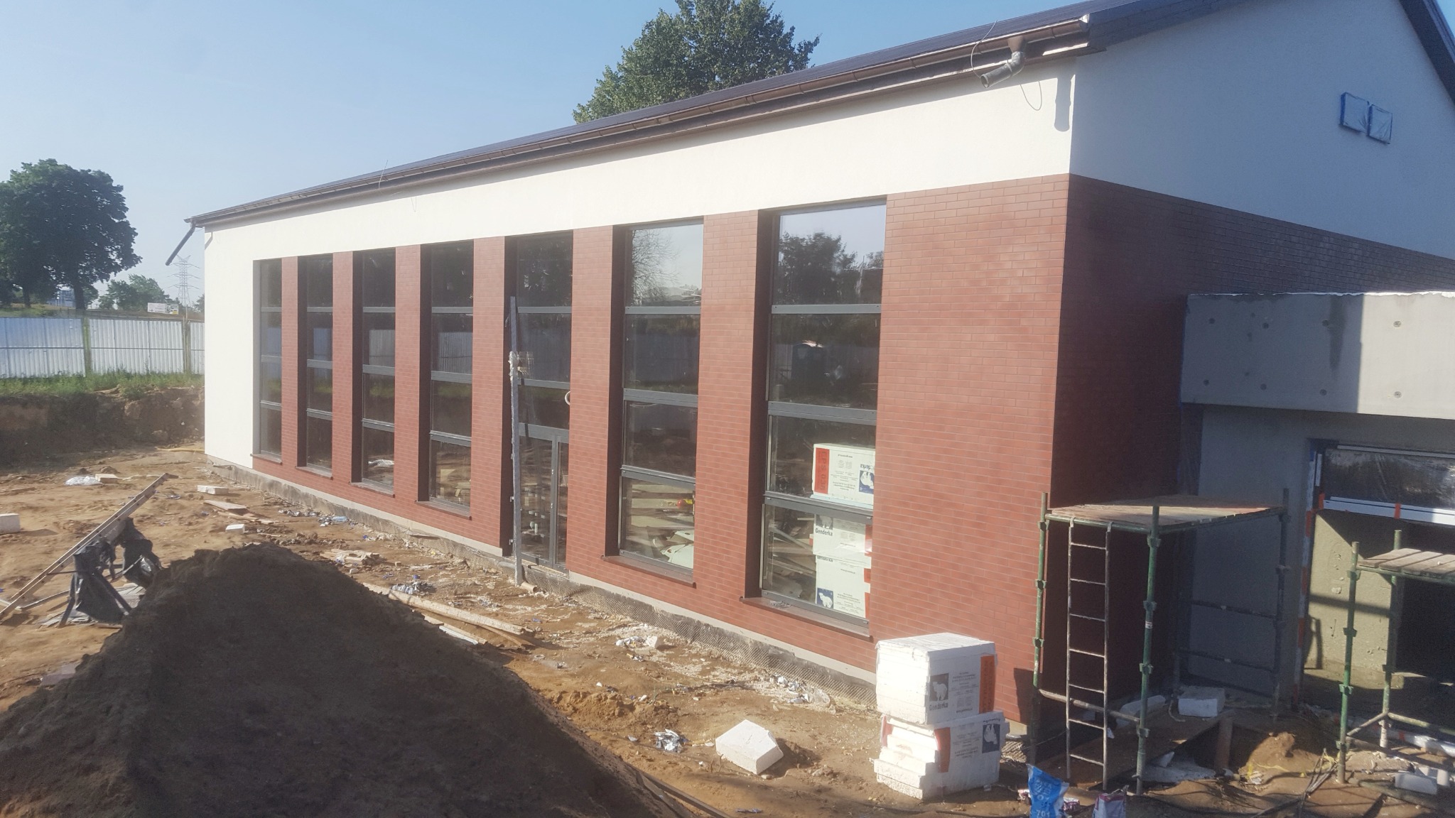 Budynek z elewacją z cegły klinkierowej w trakcie budowy, widoczne okna i rusztowania, teren wokół budynku w stanie surowym z hałdą ziemi.