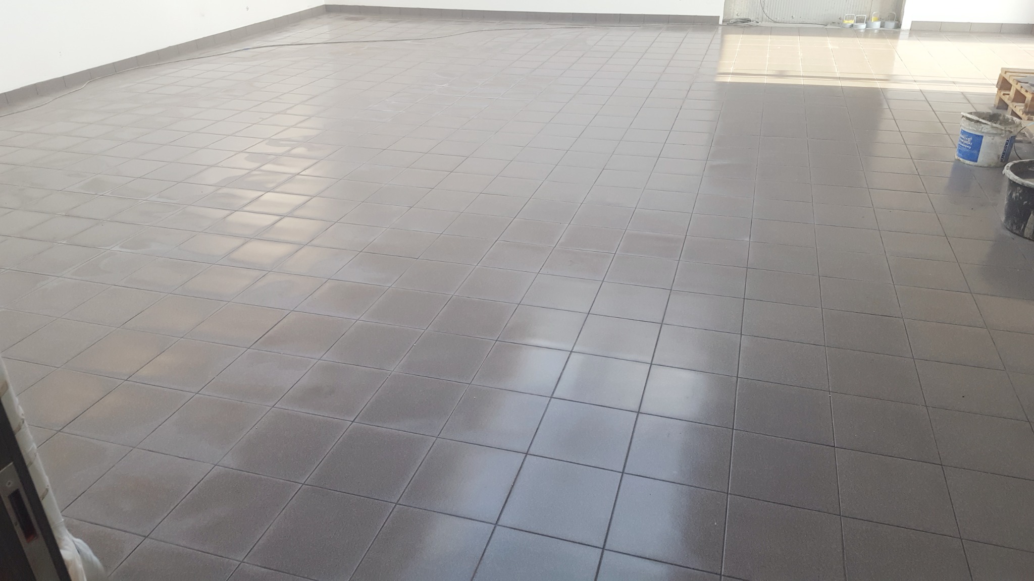 Świeżo położona, szara, kwadratowa podłoga z płytek ceramicznych w przestronnym, jasnym pomieszczeniu; widoczne wiaderka z zaprawą w prawym górnym rogu.