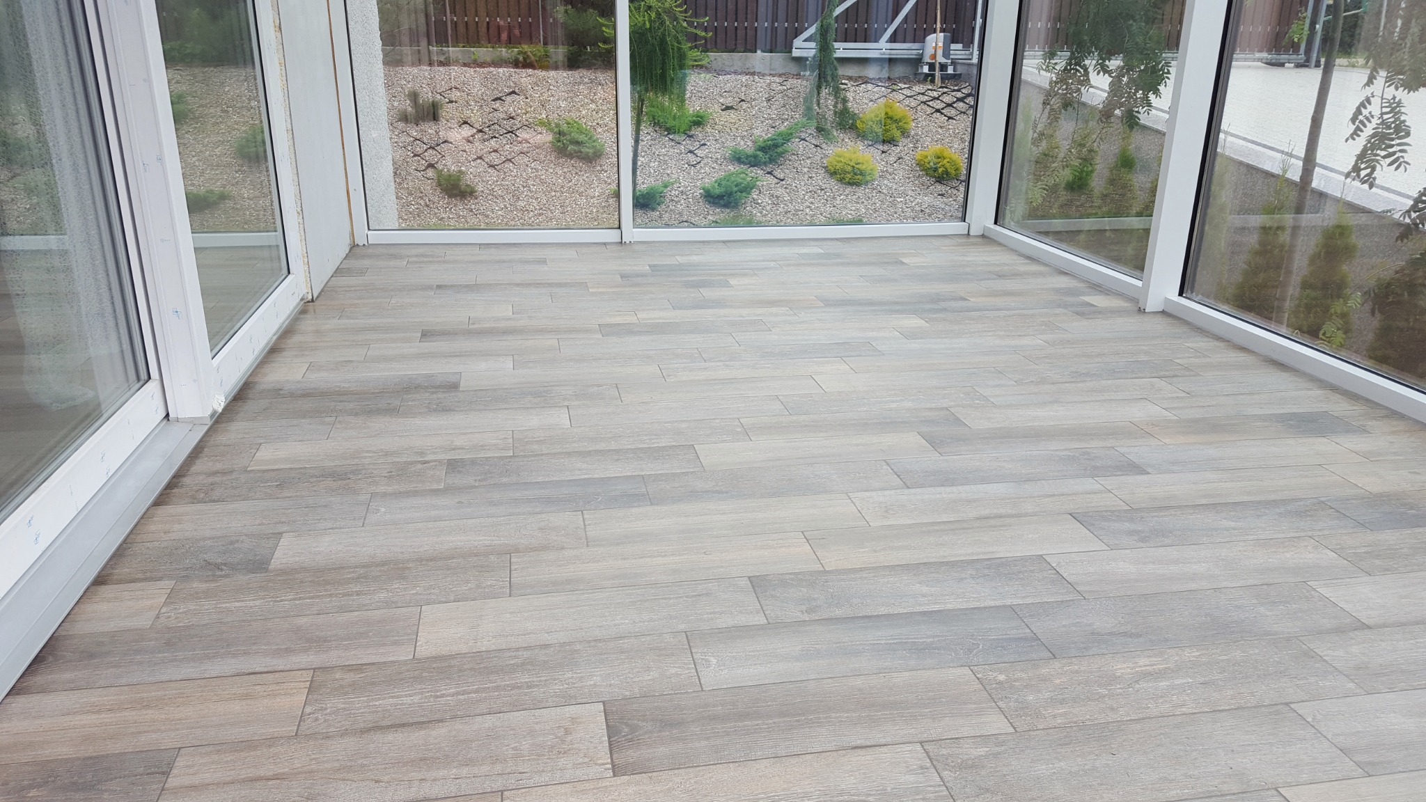 Wykończona podłoga w oranżerii, ułożona z szarych płytek imitujących drewno, widok z niskiej perspektywy w kierunku ogrodu za oknami.
