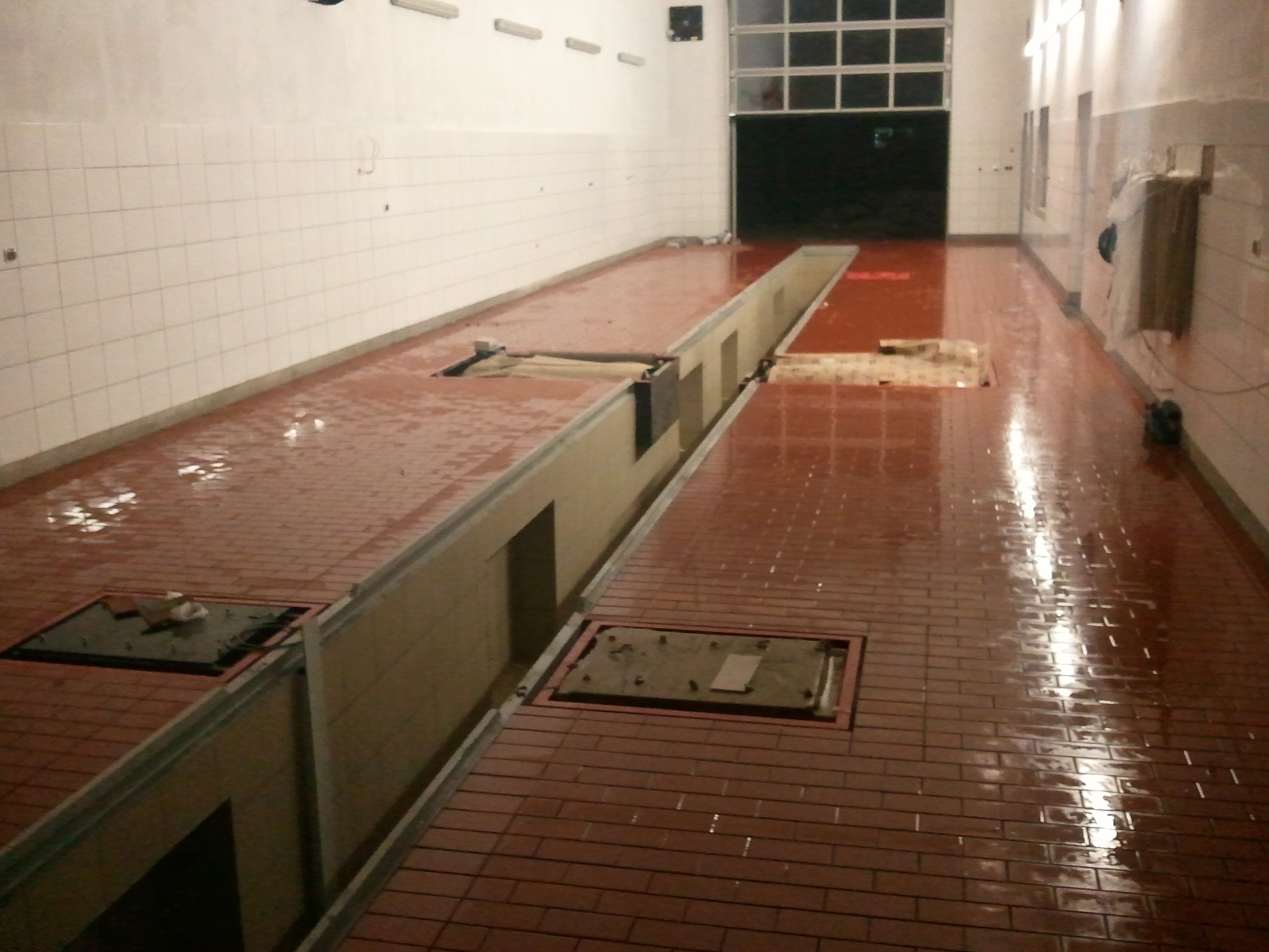 Wykończona posadzka z czerwonej terakoty z kanałem rewizyjnym i białymi płytkami na ścianach w garażu lub warsztacie samochodowym, widoczne mokre plamy na podłodze.