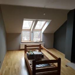 Poddasze po remoncie: ściany pomalowane w odcieniach szarości i czerni, drewniana podłoga, okna dachowe, łóżko w trakcie montażu.