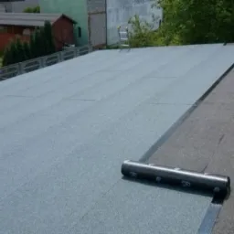 Płaski dach pokryty papą termozgrzewalną, widoczny fragment nowej warstwy oraz rolka materiału przygotowana do montażu, w tle drabina oparta o budynek.