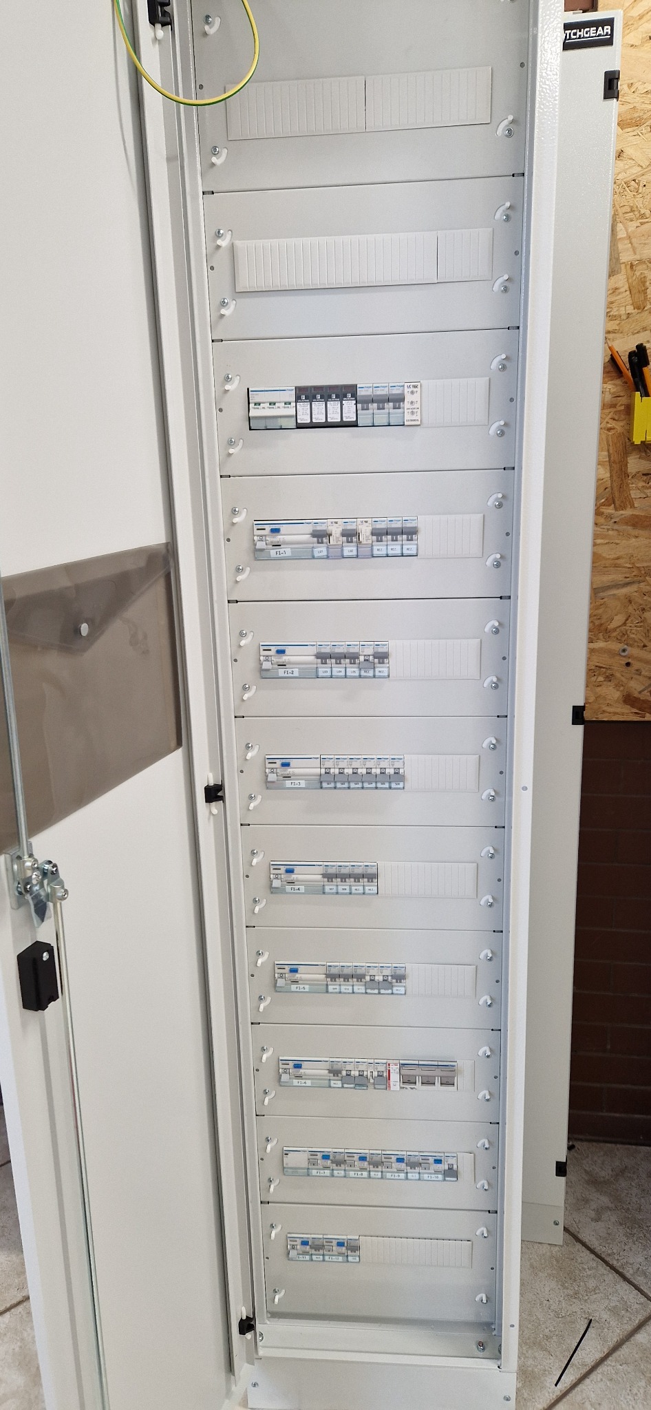 Otwarta szafa rozdzielcza z widocznymi rzędami zabezpieczeń elektrycznych i przewodami, zamontowana w pomieszczeniu z drewnianą boazerią.