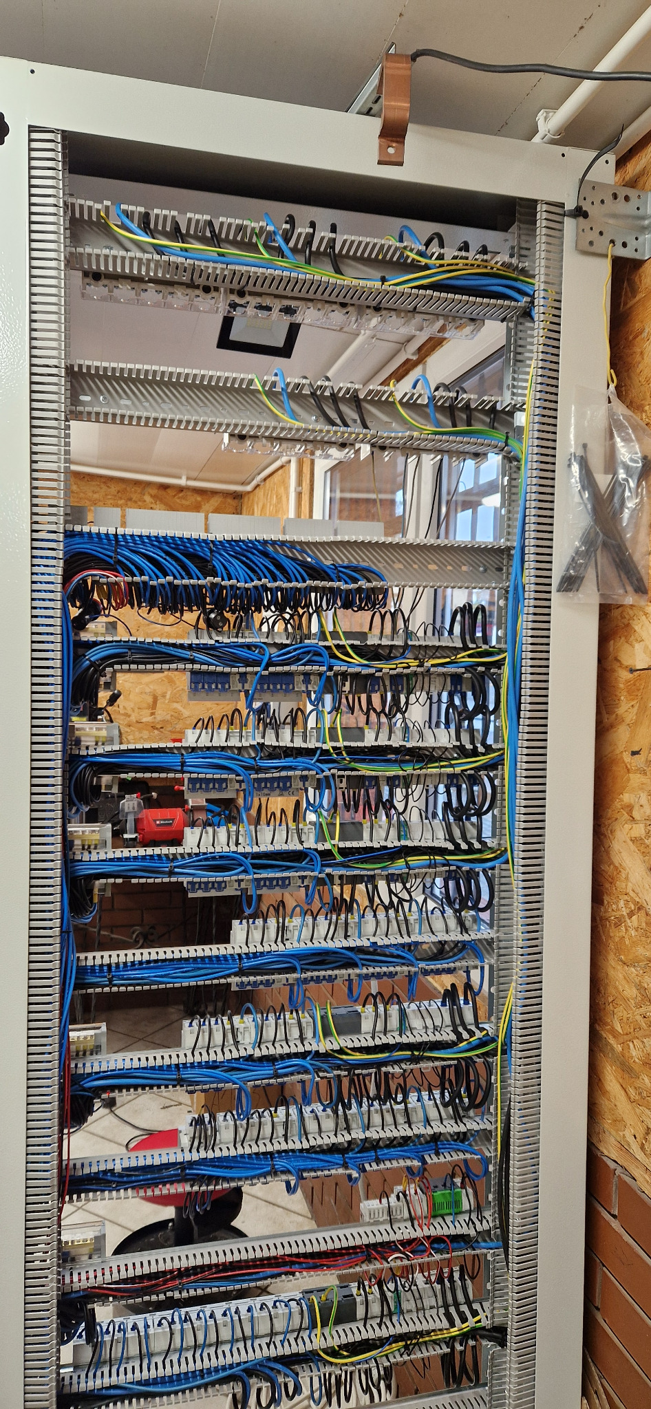 Rozdzielnica elektryczna z widocznymi przewodami w kolorach niebieskim, żółto-zielonym i czarnym, uporządkowanymi na listwach montażowych wewnątrz szafy, z drewnianą ścianą w tle.