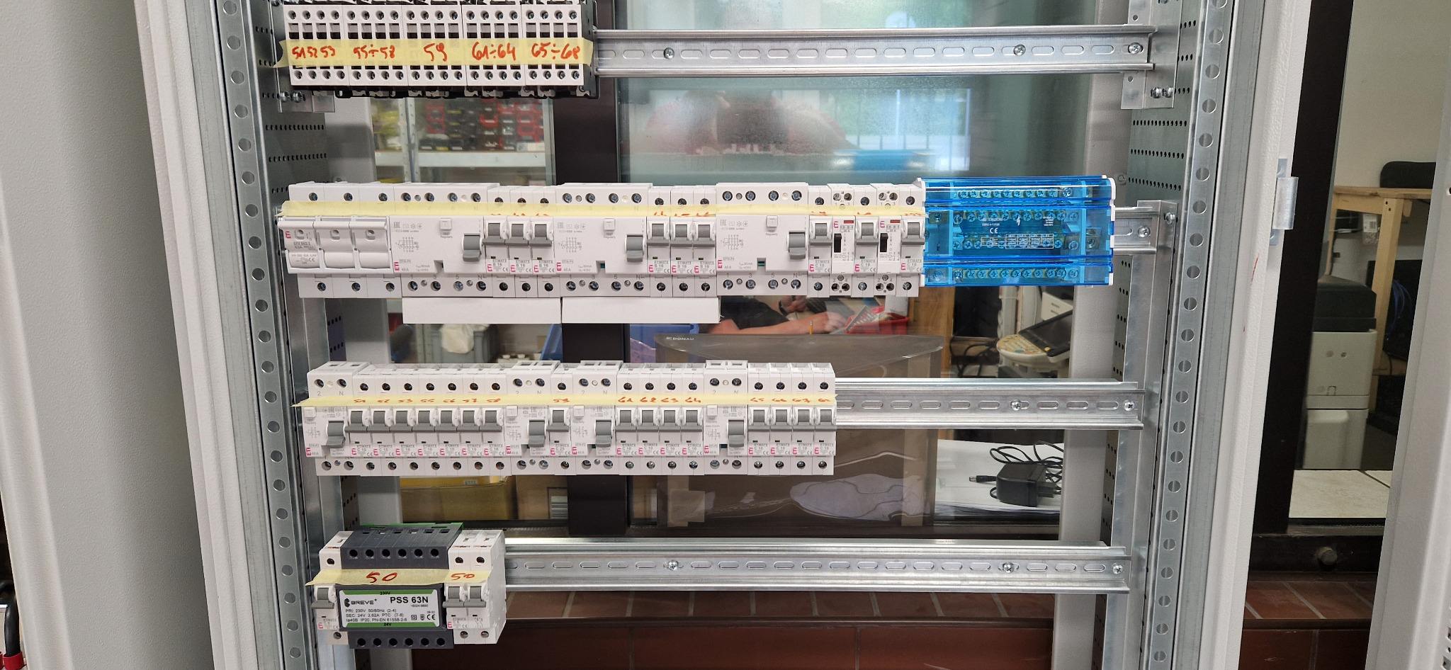 Rozdzielnica elektryczna z zamontowanymi bezpiecznikami i listwami zaciskowymi, widoczne oznaczenia numerów na taśmie, na drugim planie elementy wyposażenia pomieszczenia.
