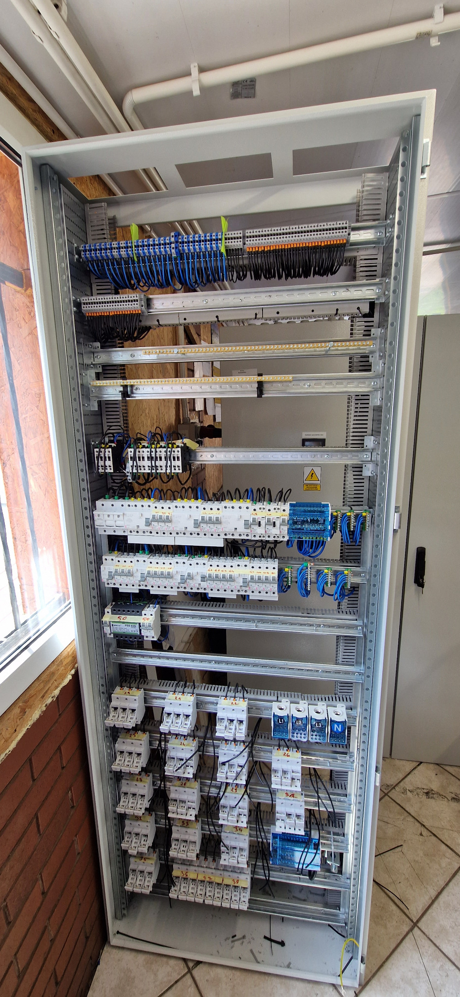 Otwarta szafa sterownicza z zamontowanymi bezpiecznikami, przekaźnikami i okablowaniem w kolorach niebieskim i czarnym, widok z boku z elementami ściany z cegły w tle.