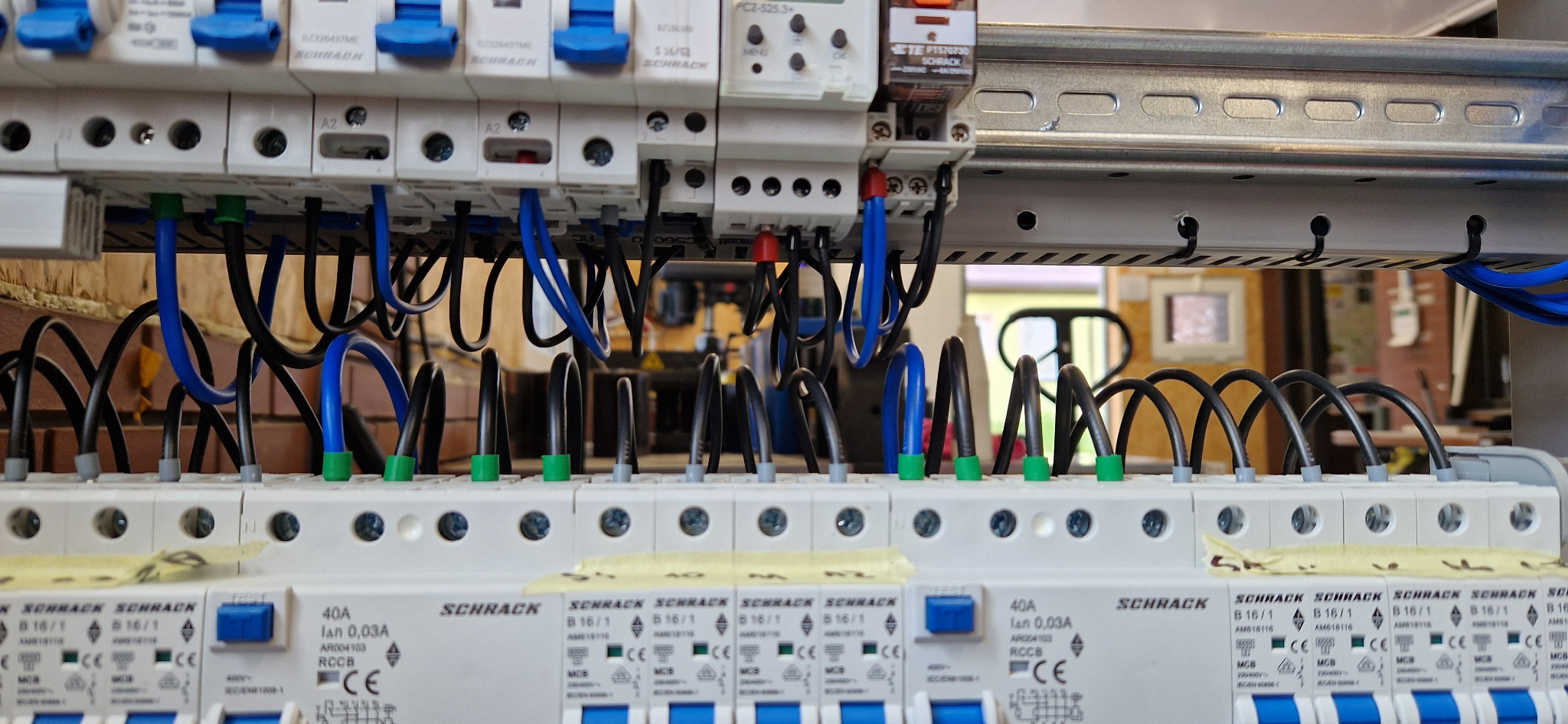 Szczegółowe ujęcie tablicy rozdzielczej z bezpiecznikami Schrack, widoczne przewody w kolorach czarnym i niebieskim, z zielonymi i szarymi końcówkami, oraz metalowa szyna montażowa