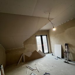 GRUBE WAŁKI SPÓŁKA Z OGRANICZONĄ ODPOWIEDZIALNOŚCIĄ - Poddasze z oknem, w trakcie montażu płyt karton-gips na skośnym suficie. Widoczne narzędzia i materiały budowlane. Ściany przygotowane pod malowanie.