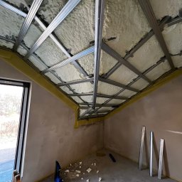 GRUBE WAŁKI SPÓŁKA Z OGRANICZONĄ ODPOWIEDZIALNOŚCIĄ - Poddasze z ociepleniem pianą i metalową konstrukcją pod płytę G-K, widok z dołu. Na podłodze resztki piany i narzędzia. Okno w lewym rogu.