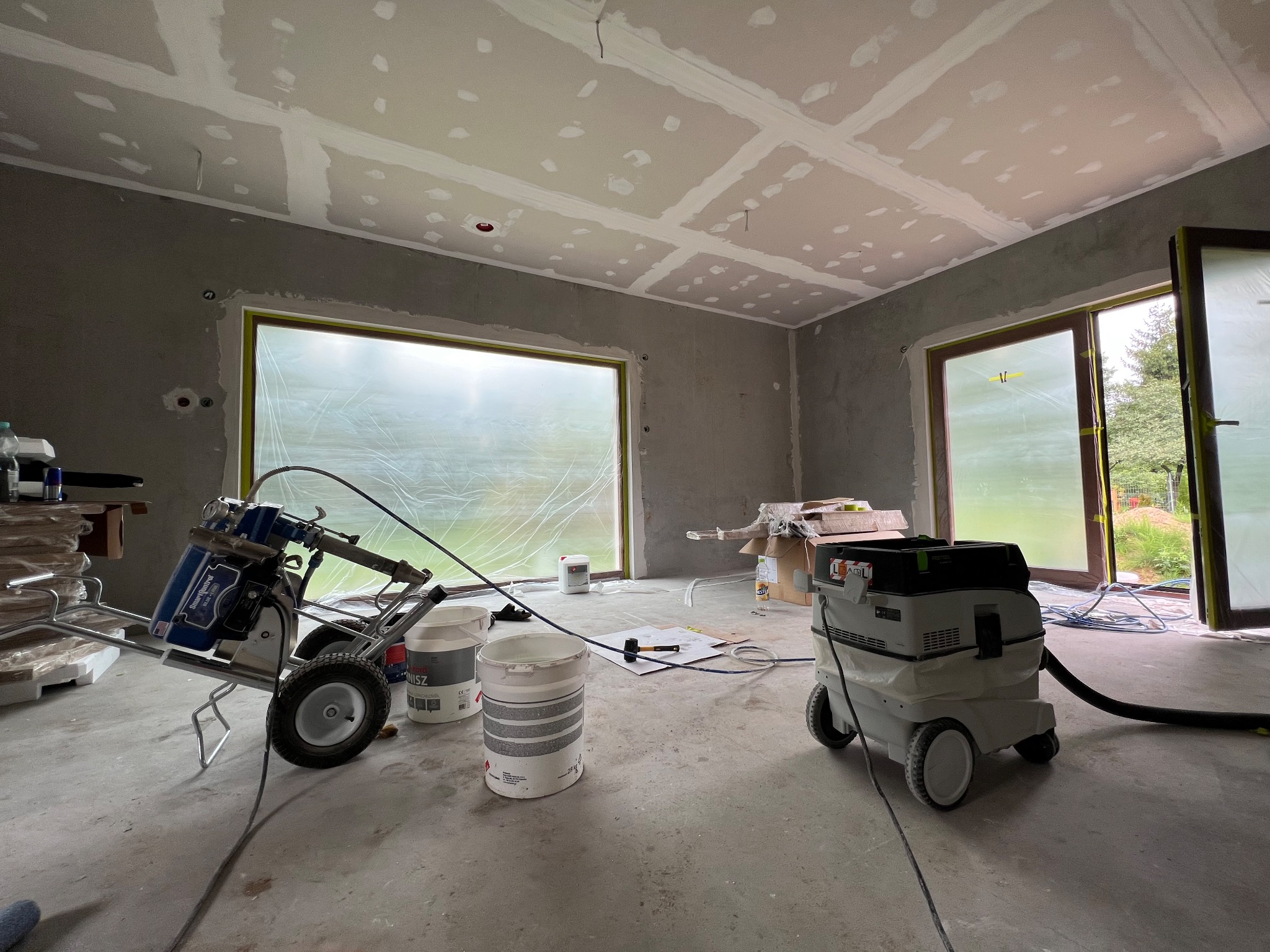 Wnętrze w trakcie prac wykończeniowych: ściany przygotowane pod malowanie, sufit z płyt kartonowo-gipsowych, sprzęt malarski i odkurzacz przemysłowy. Okna zabezpieczone folią.