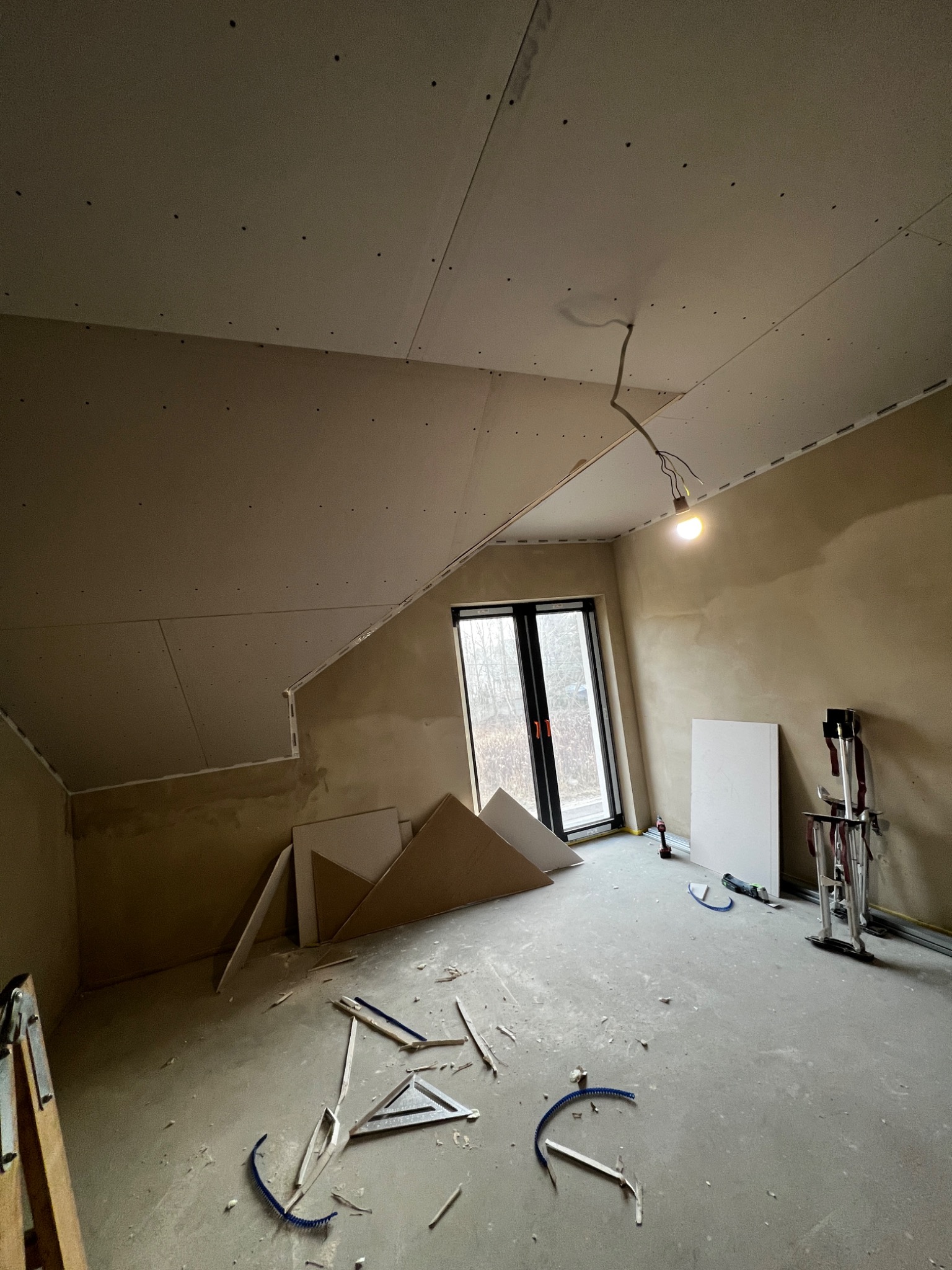 Poddasze z oknem, w trakcie montażu płyt karton-gips na skośnym suficie. Widoczne narzędzia i materiały budowlane. Ściany przygotowane pod malowanie.