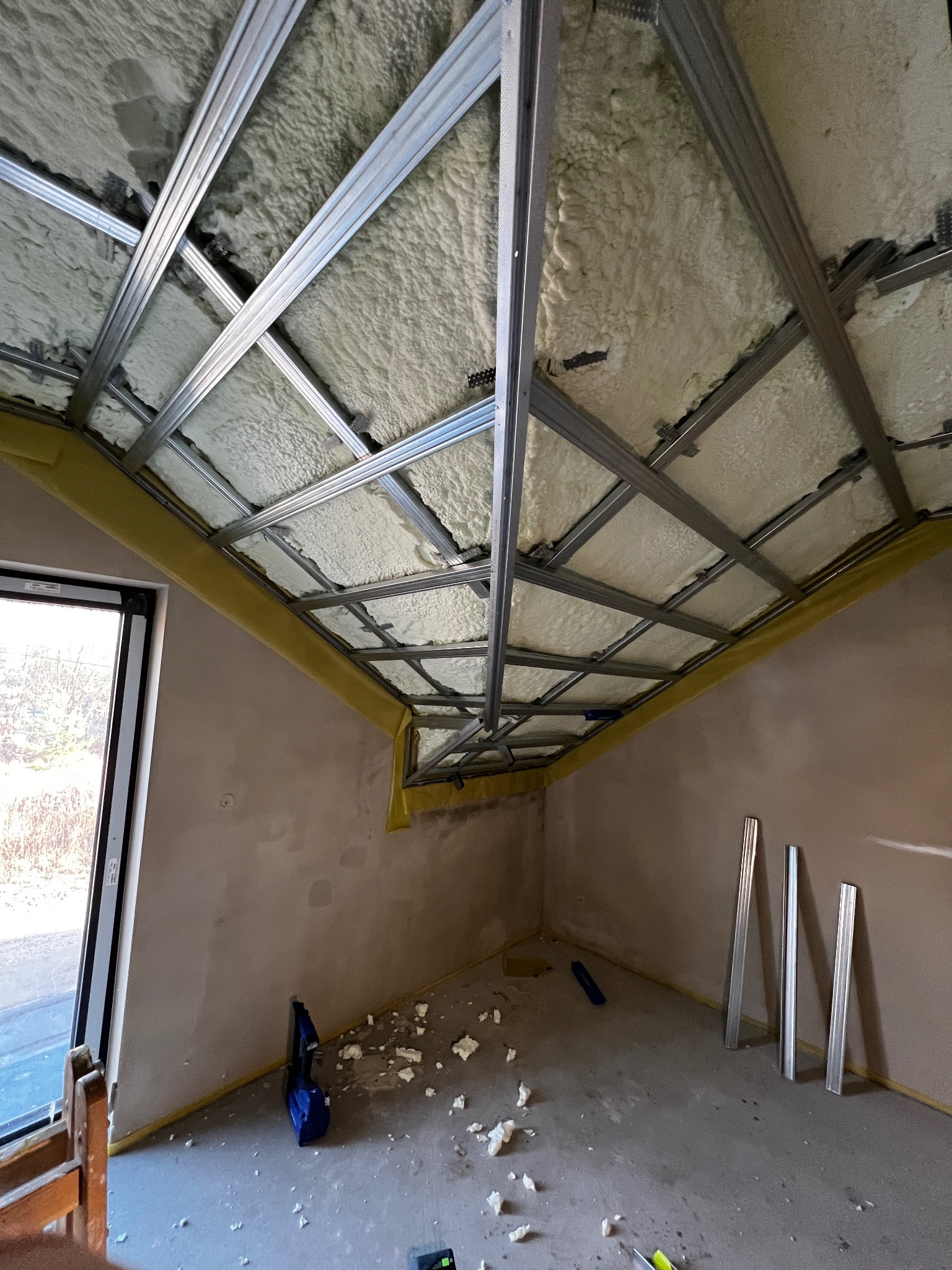 Poddasze z ociepleniem pianą i metalową konstrukcją pod płytę G-K, widok z dołu. Na podłodze resztki piany i narzędzia. Okno w lewym rogu.