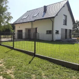 Nowoczesny dom jednorodzinny z szarą dachówką, otoczony metalowym ogrodzeniem panelowym na betonowej podmurówce, z zielonym trawnikiem na pierwszym planie.