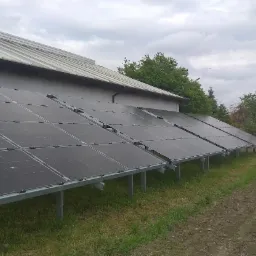 Rząd paneli fotowoltaicznych zamontowanych na metalowej konstrukcji, przylegający do ściany budynku gospodarczego z blachodachówką, na tle pochmurnego nieba i pola uprawnego z widocznym kamieniem.