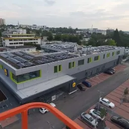 Widok z góry na budynek sklepu z panelami słonecznymi na dachu, parkingiem i otaczającą zabudową miejską w tle.