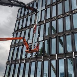 WB-Fassaden - Montaż paneli elewacyjnych na wysokim biurowcu z użyciem podnośnika koszowego i dźwigu, widoczne elementy konstrukcji budynku oraz pracownicy w kaskach.